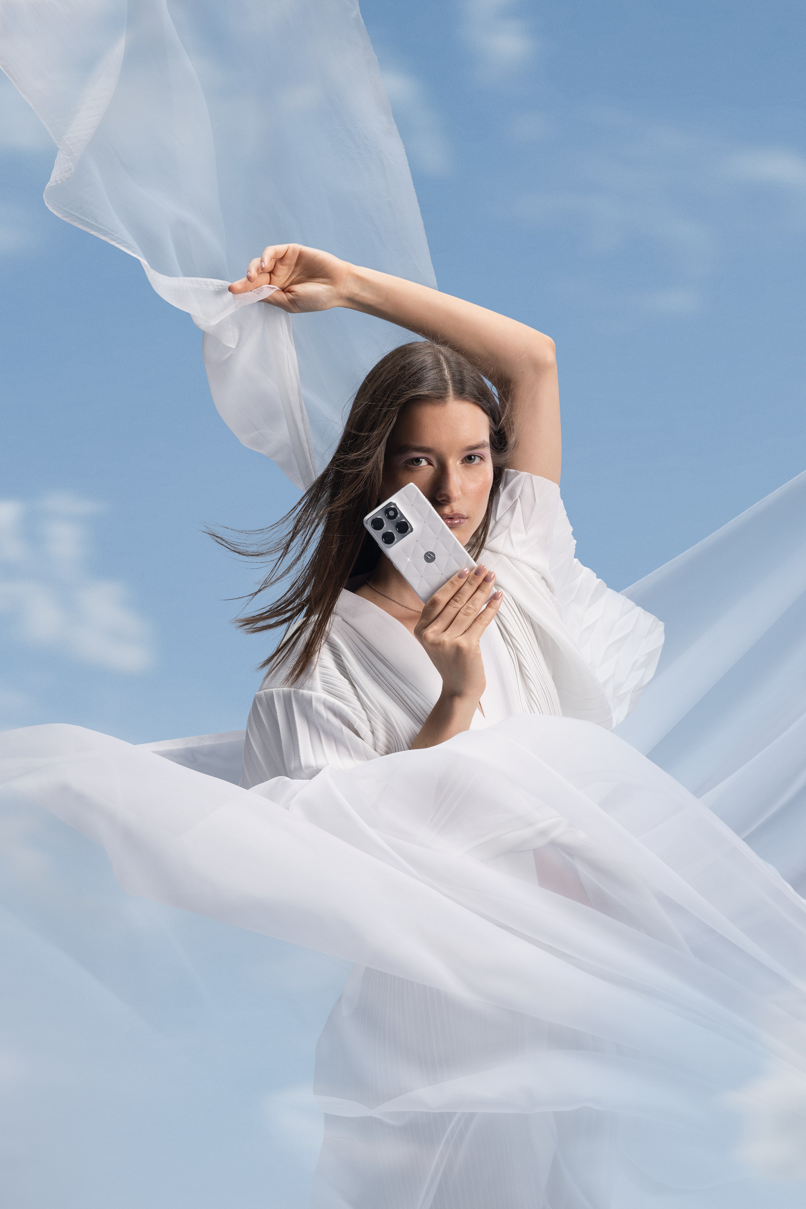 Woman lifts sheer white fabric overhead and holds a white phone beside her face against clouds