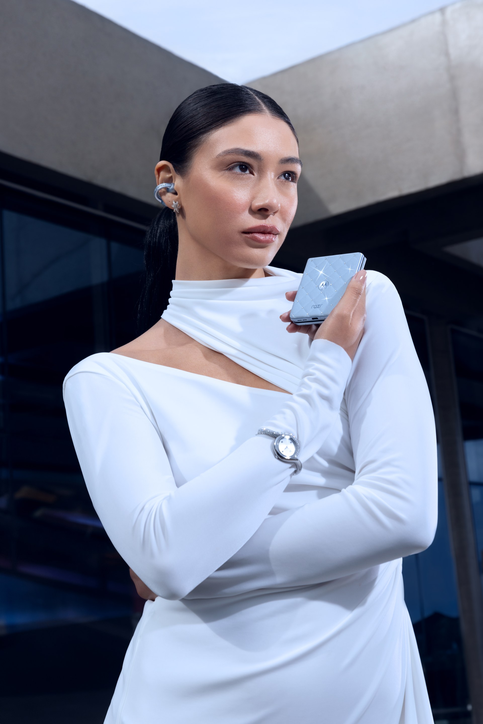 Woman in a white cut-out top with a crystal ear cuff holds a pale blue phone in front of glass architecture
