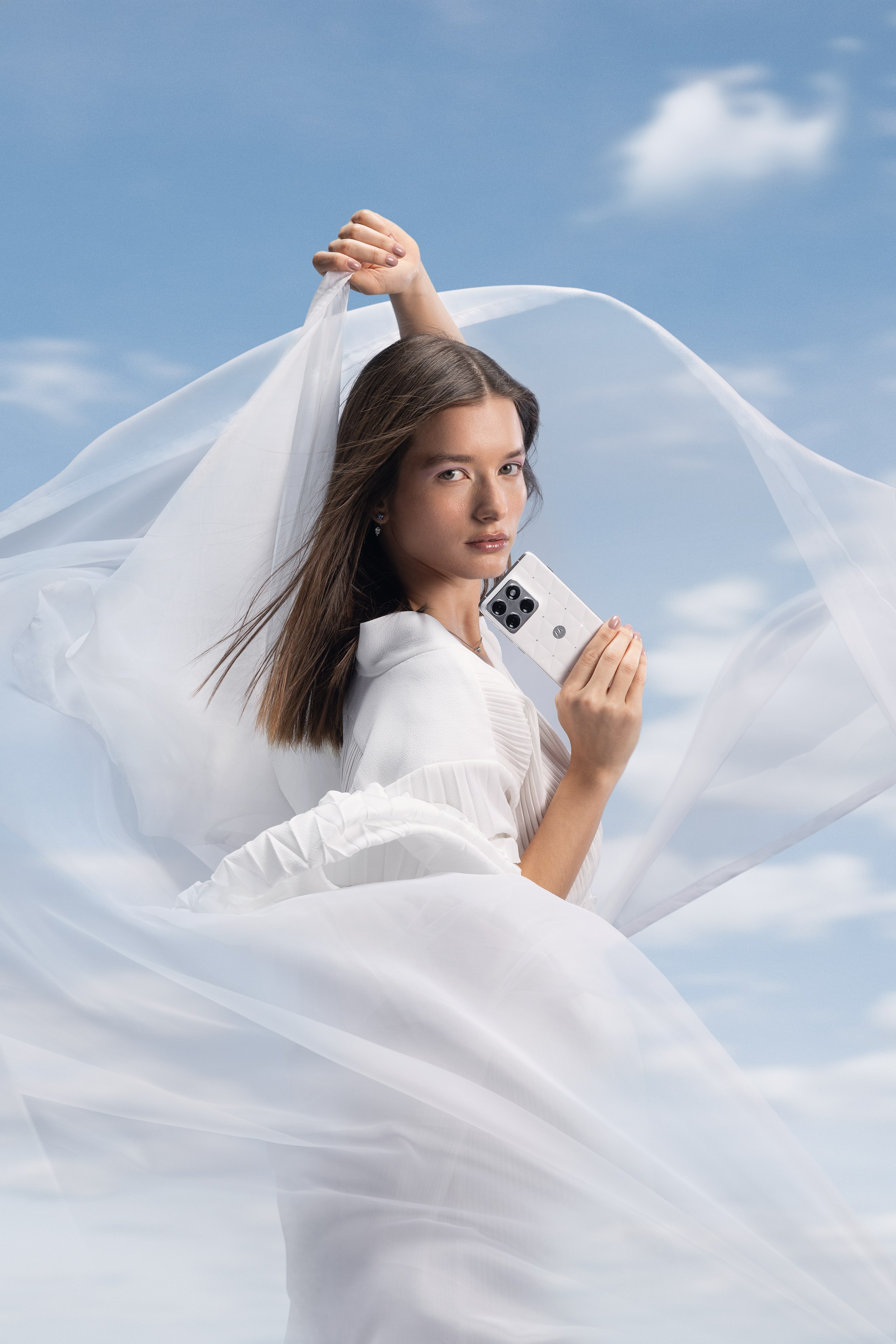 Woman in white holds up a white phone as sheer white fabric swirls around her against a blue sky