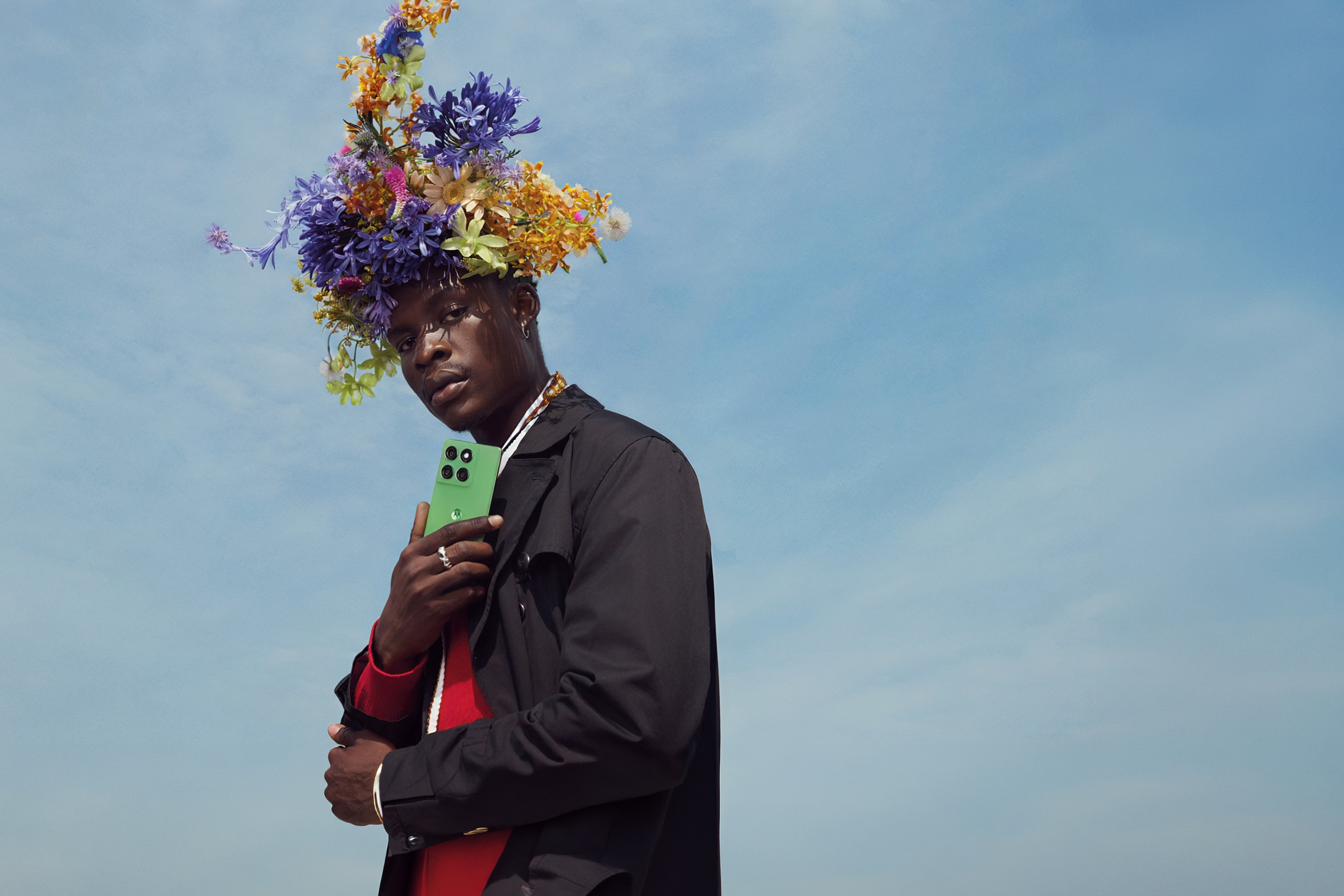 Low-angle portrait of a man in a floral headdress and black jacket holding a green phone, sky behind