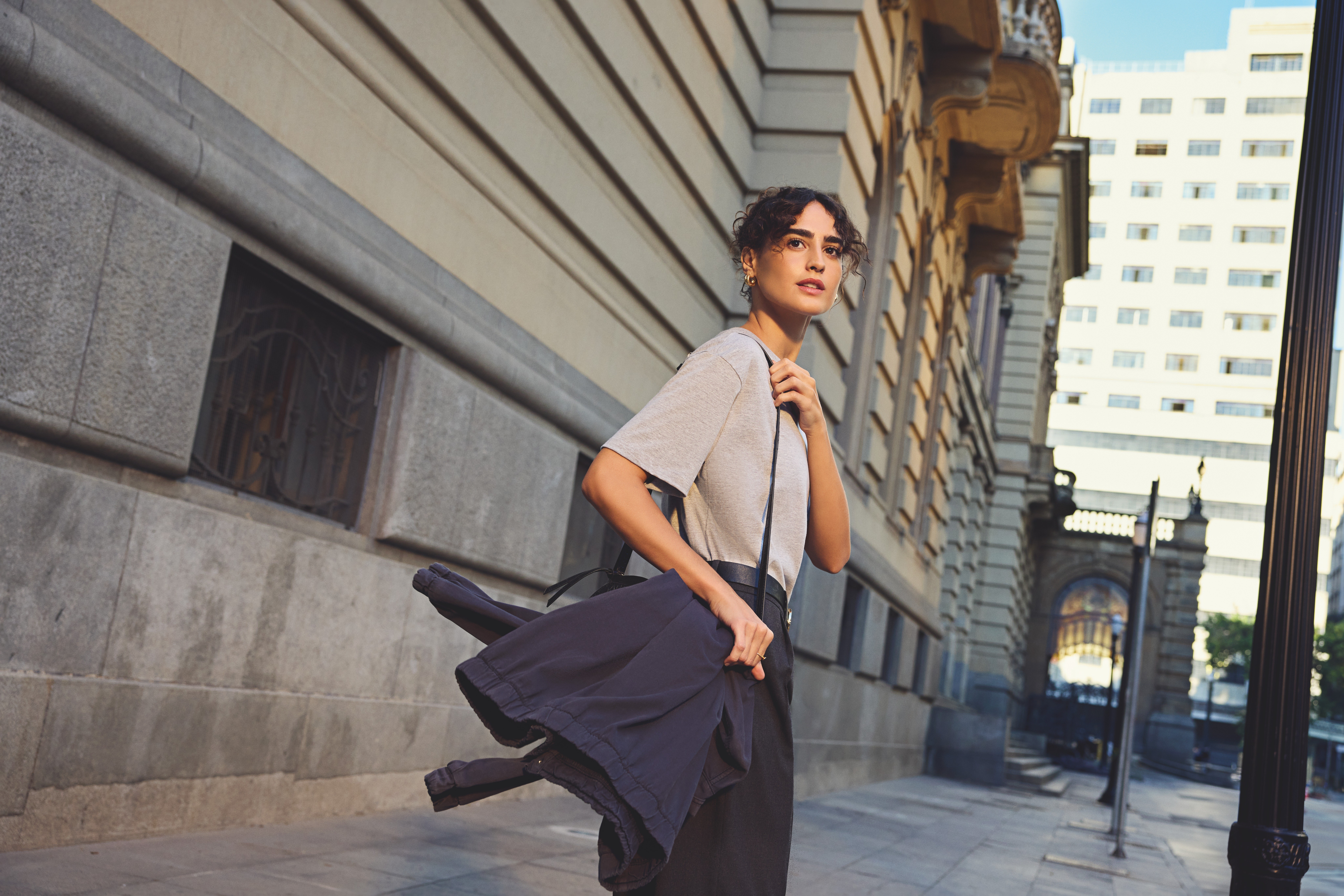 Woman with curly hair and a grey tee walks past an ornate stone building, a navy jacket swinging from her hand, city towers behind