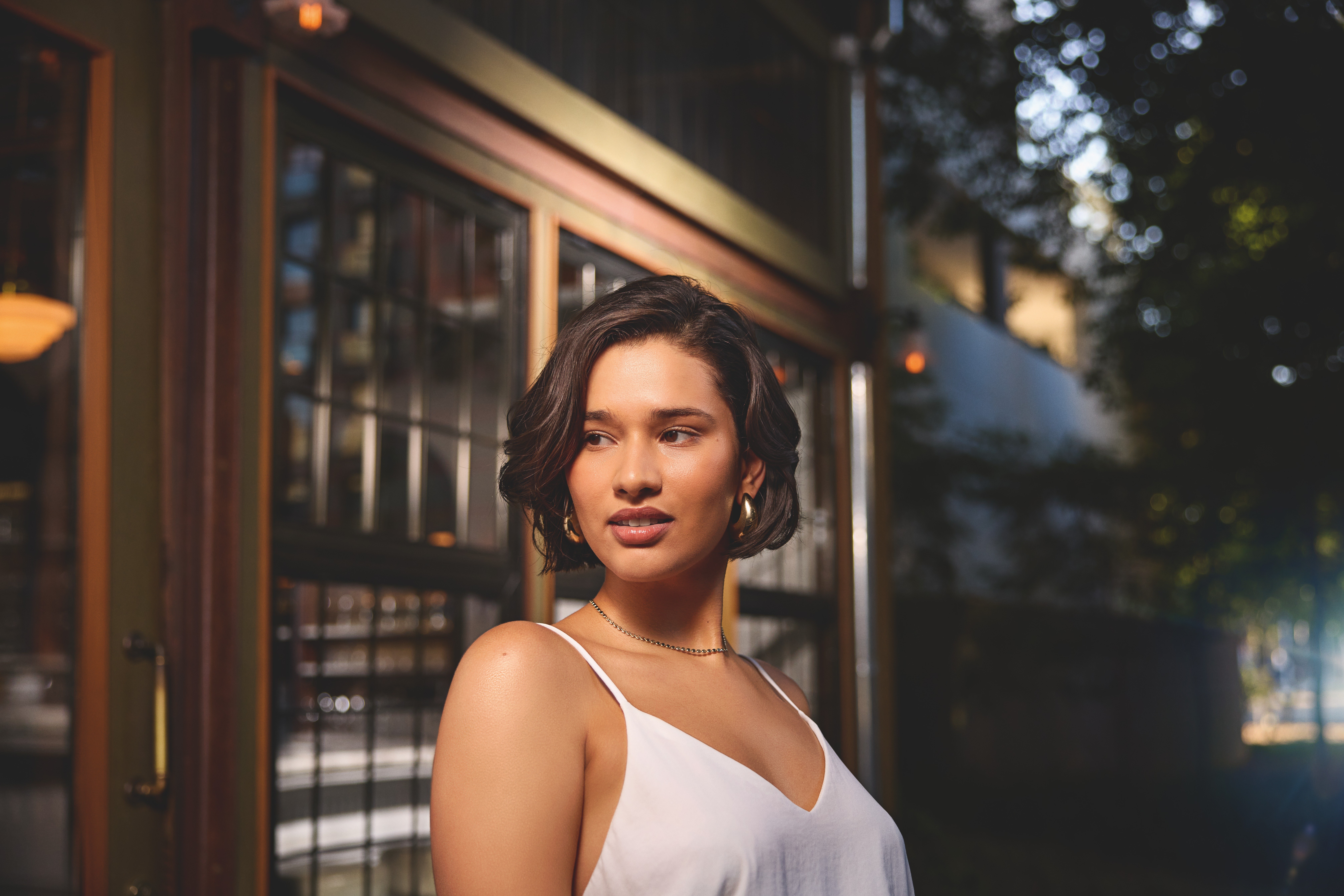 Portrait of a woman with a short wavy bob, gold earrings and a white camisole, glancing aside outside a glass-paneled cafe at dusk