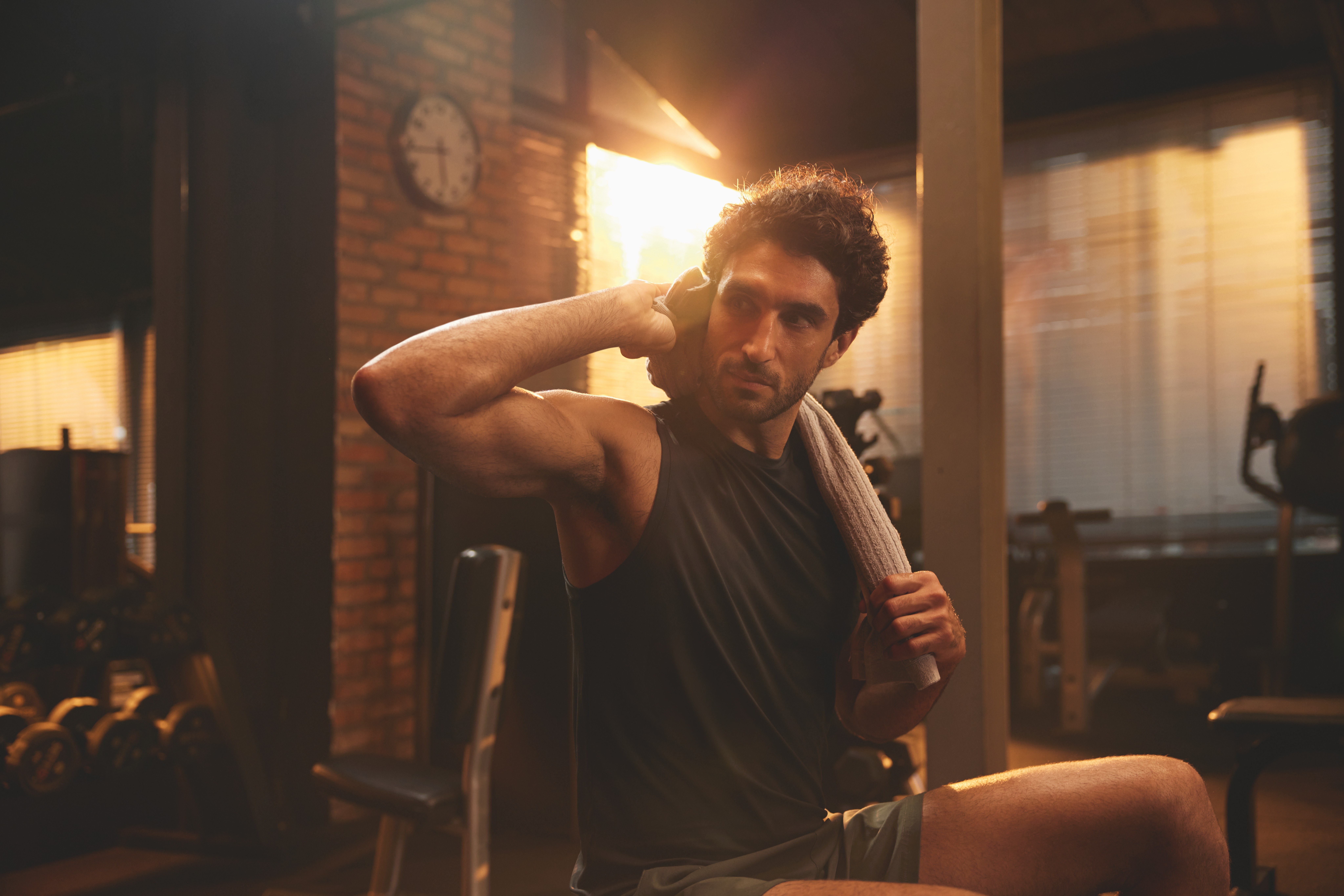 Man in a black tank top wipes his neck with a towel on a gym bench, warm sunlight flaring through the window behind him