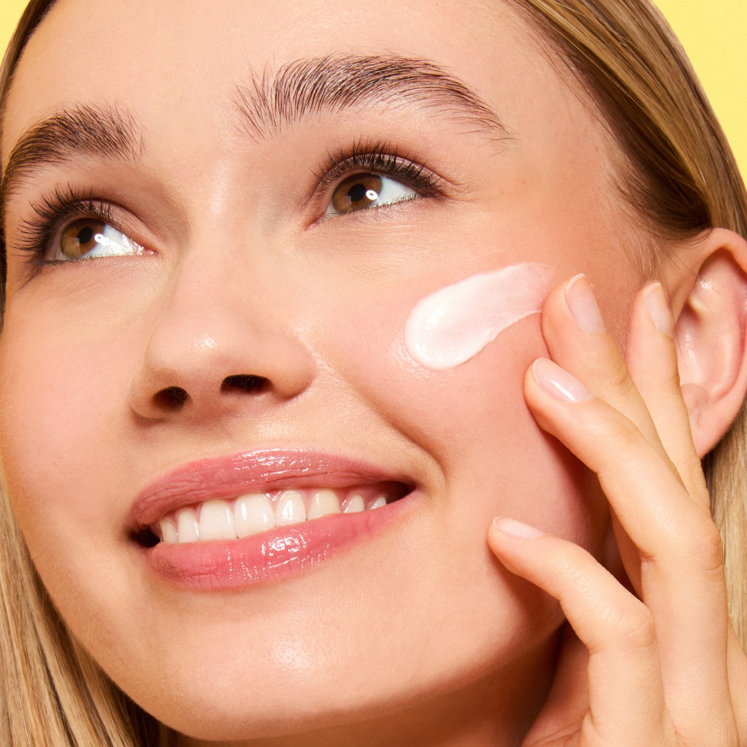 Close-up of a smiling woman looking upward as she smooths a swipe of white cream onto her cheek against a yellow backdrop