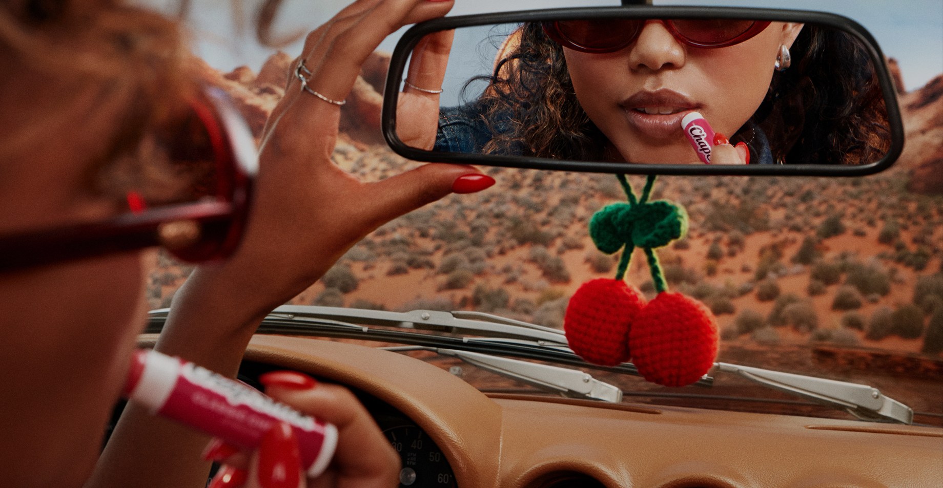 Woman in red sunglasses applies ChapStick in a convertible's rearview mirror as she drives through desert, crochet cherries hanging below