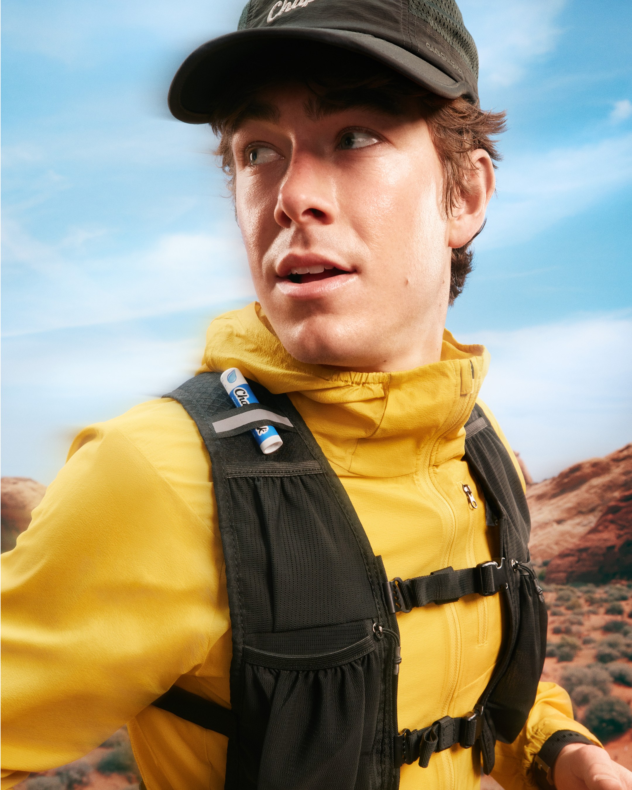 Runner in a yellow jacket and cap with a ChapStick tube clipped to his hydration vest strap, red desert rock behind him