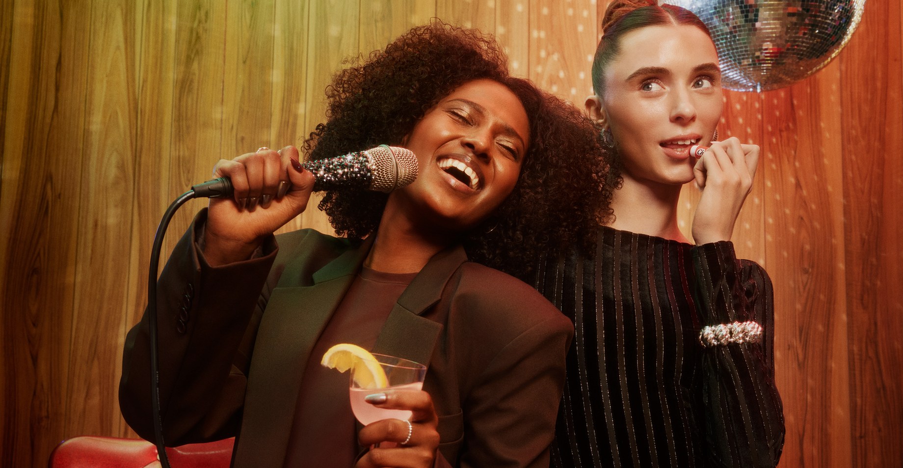 Two women at a wood-paneled karaoke night, one singing into a sparkly microphone with a cocktail, the other applying lip balm
