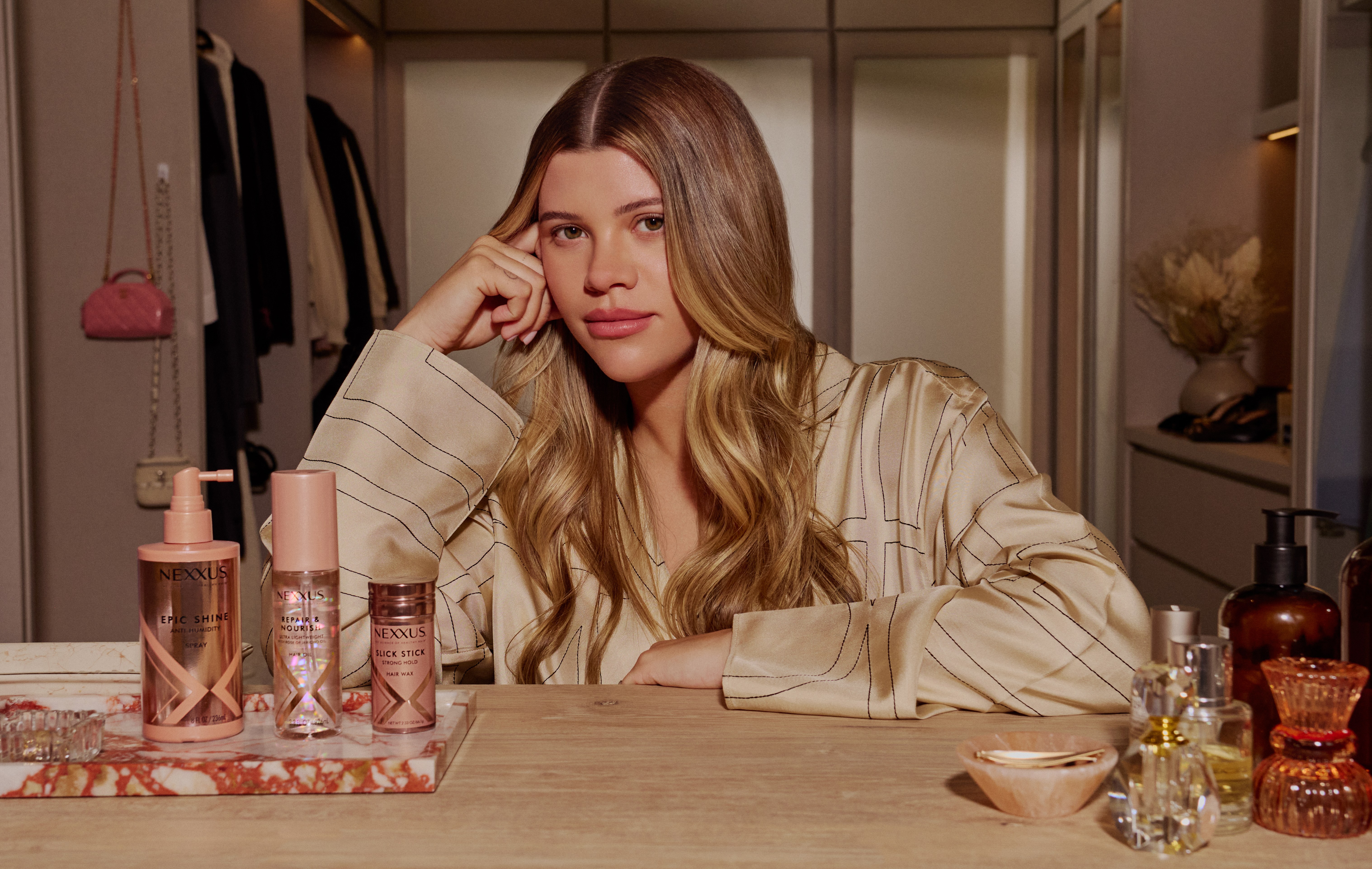 Woman with long wavy hair rests her head on her hand at a wooden vanity in a walk-in closet, Nexxus styling products lined up beside her