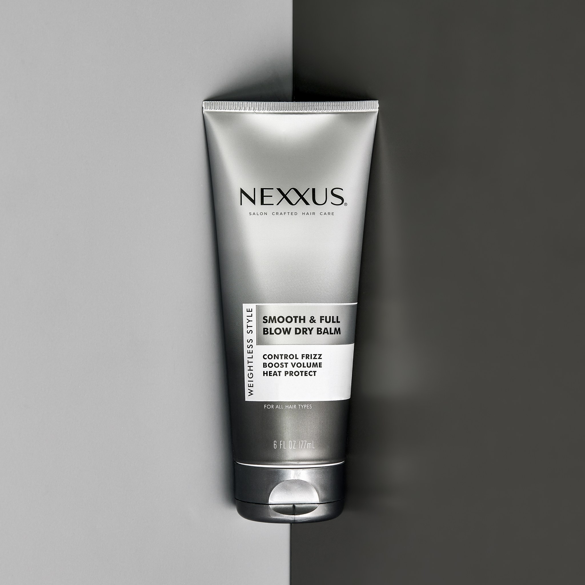 Product shot of a silver Nexxus Smooth & Full Blow Dry Balm tube set across a split grey and charcoal background
