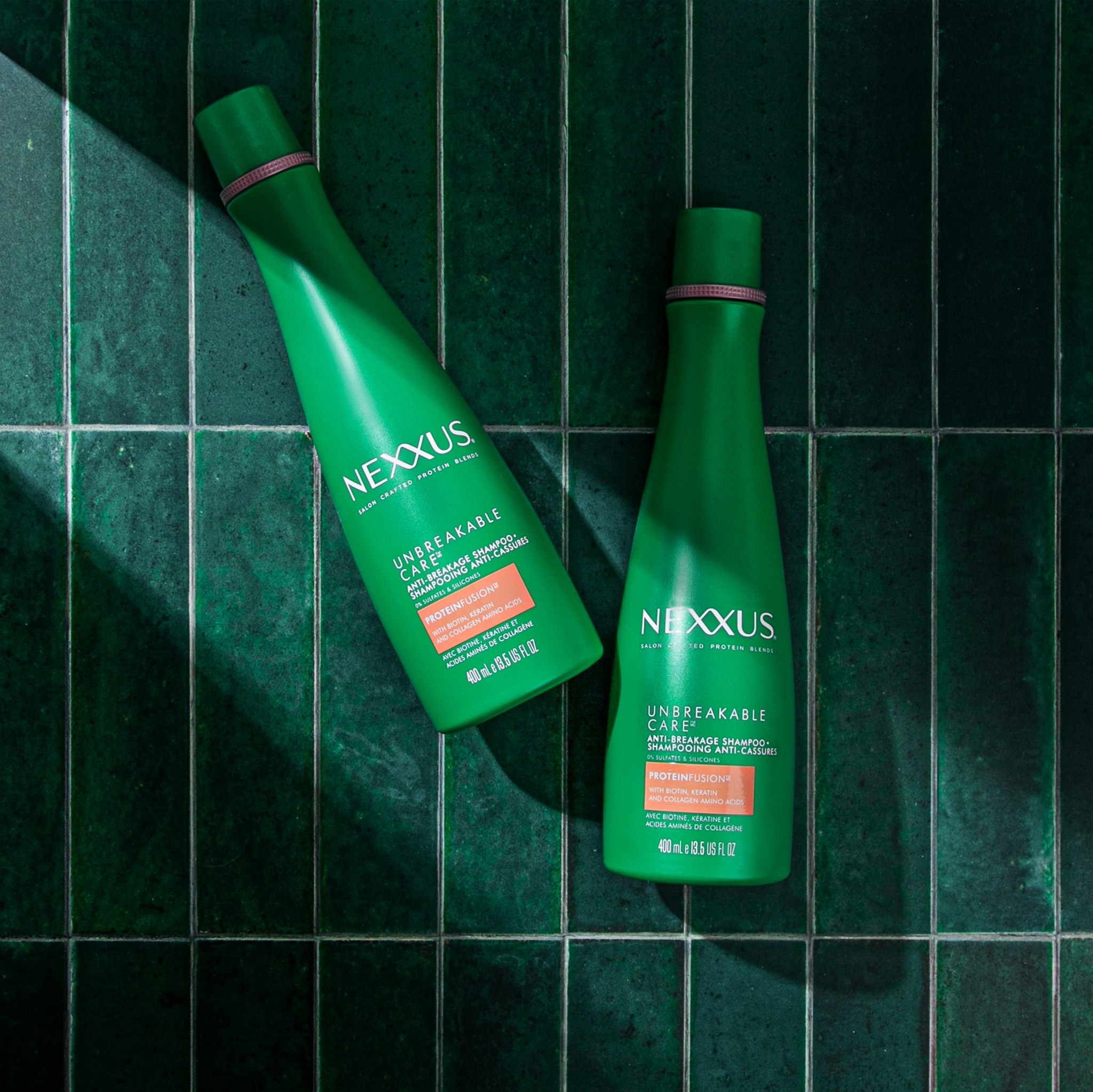 Two green Nexxus Unbreakable Care anti-breakage shampoo bottles lying on dark green glazed tiles in hard sunlight