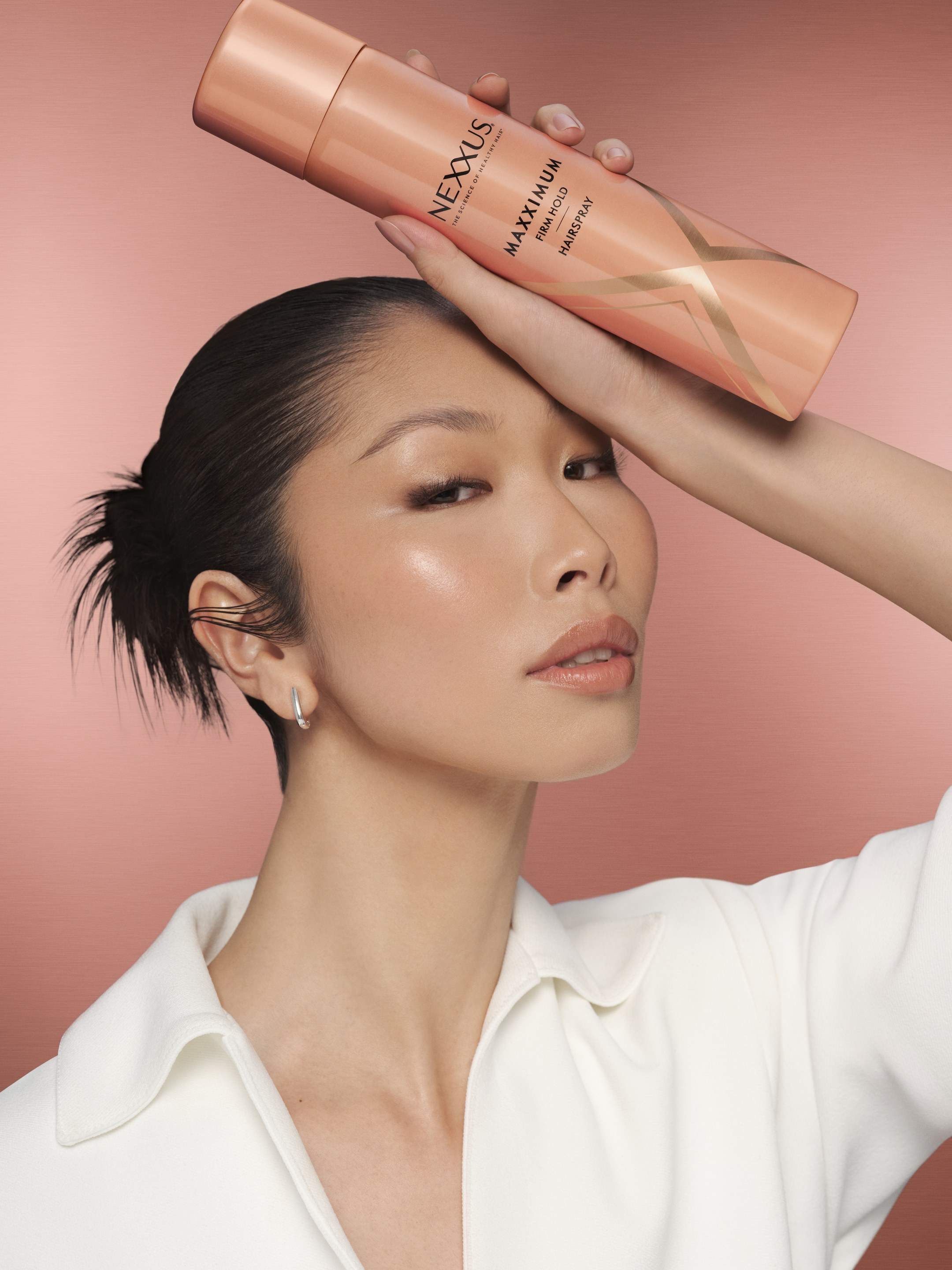 Model with slicked-back hair holds a pink Nexxus bottle above her head against a pink backdrop
