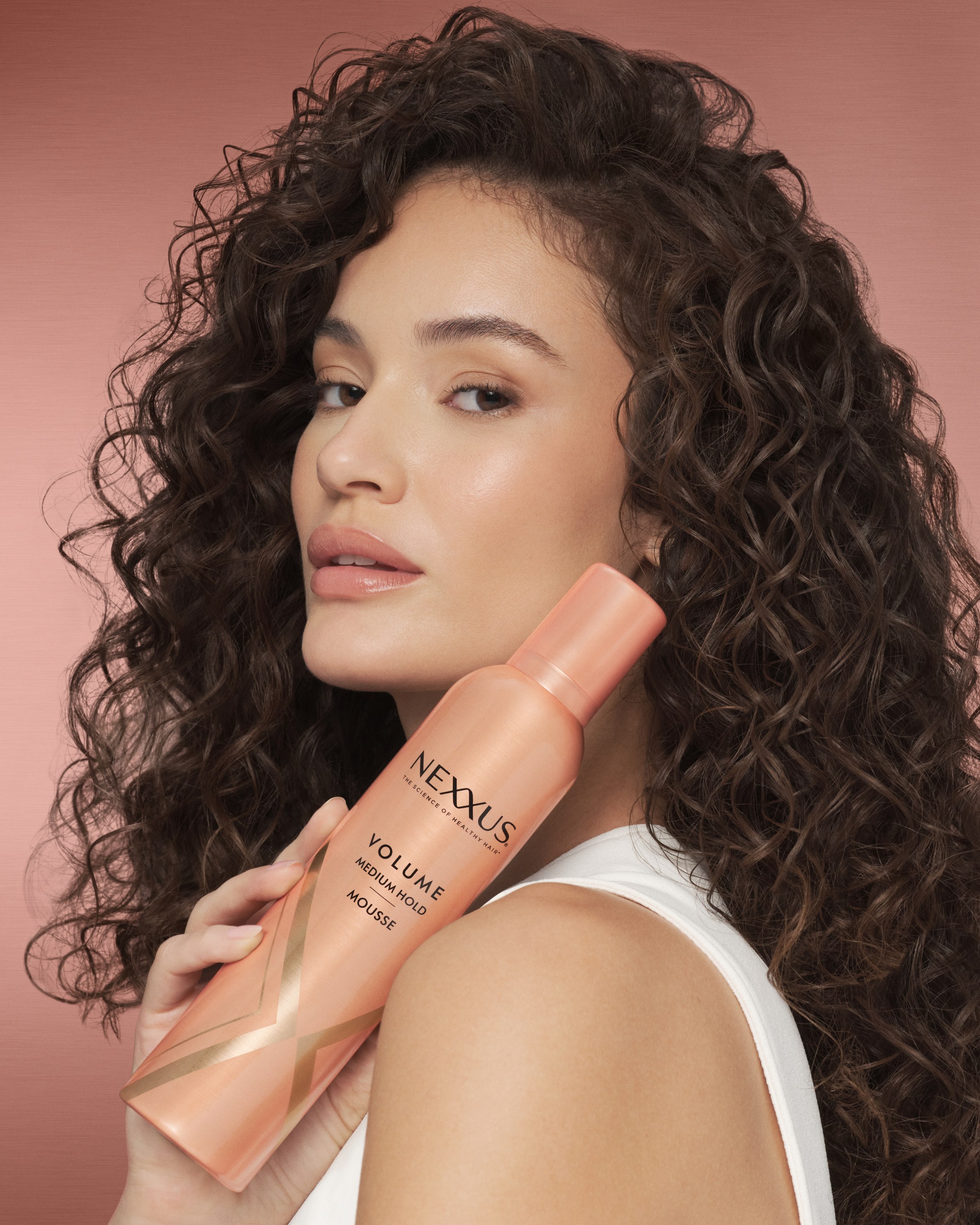 Woman with voluminous dark curls rests a pink Nexxus bottle on her shoulder against a pink backdrop