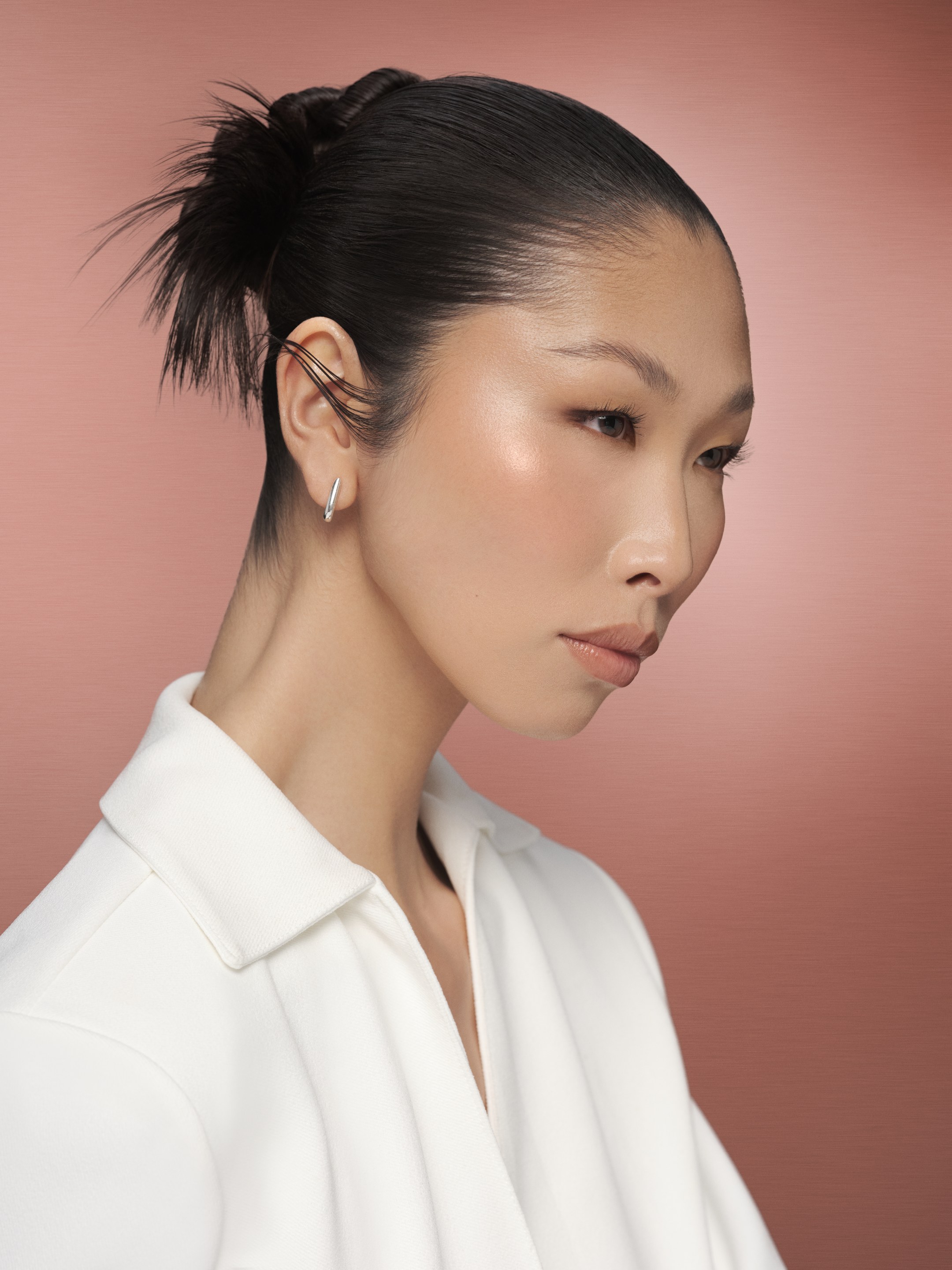 Profile of a model with a sleek bun and a white shirt against a dusty pink backdrop