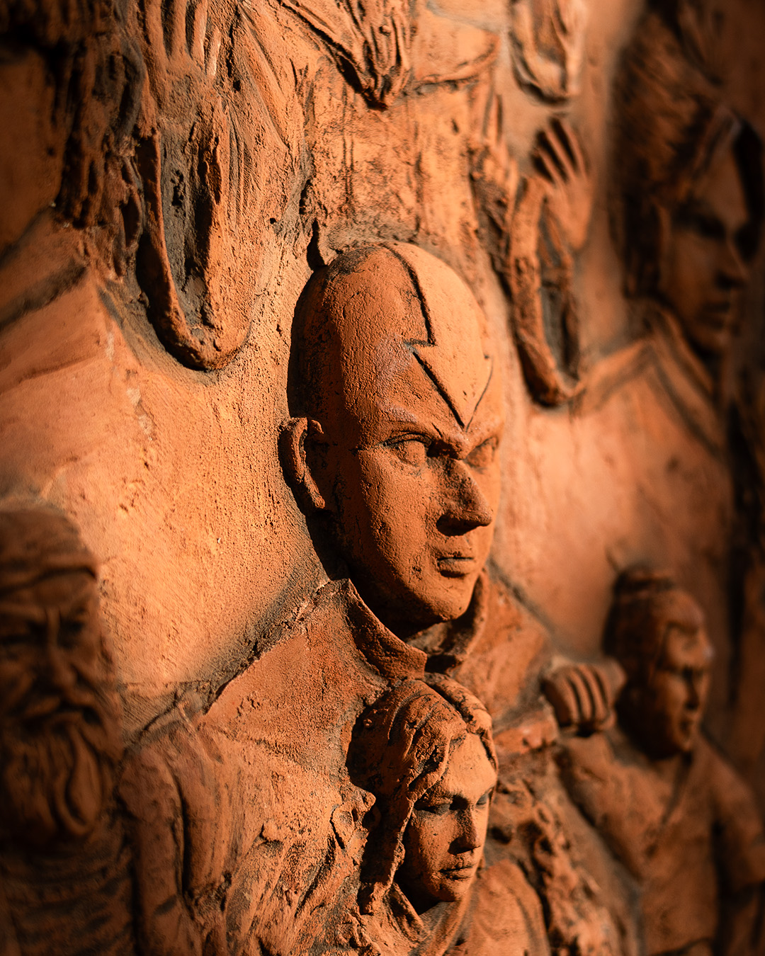 Close-up of a clay relief sculpture: the carved face of a bald boy with an arrow mark on his head, among other figures