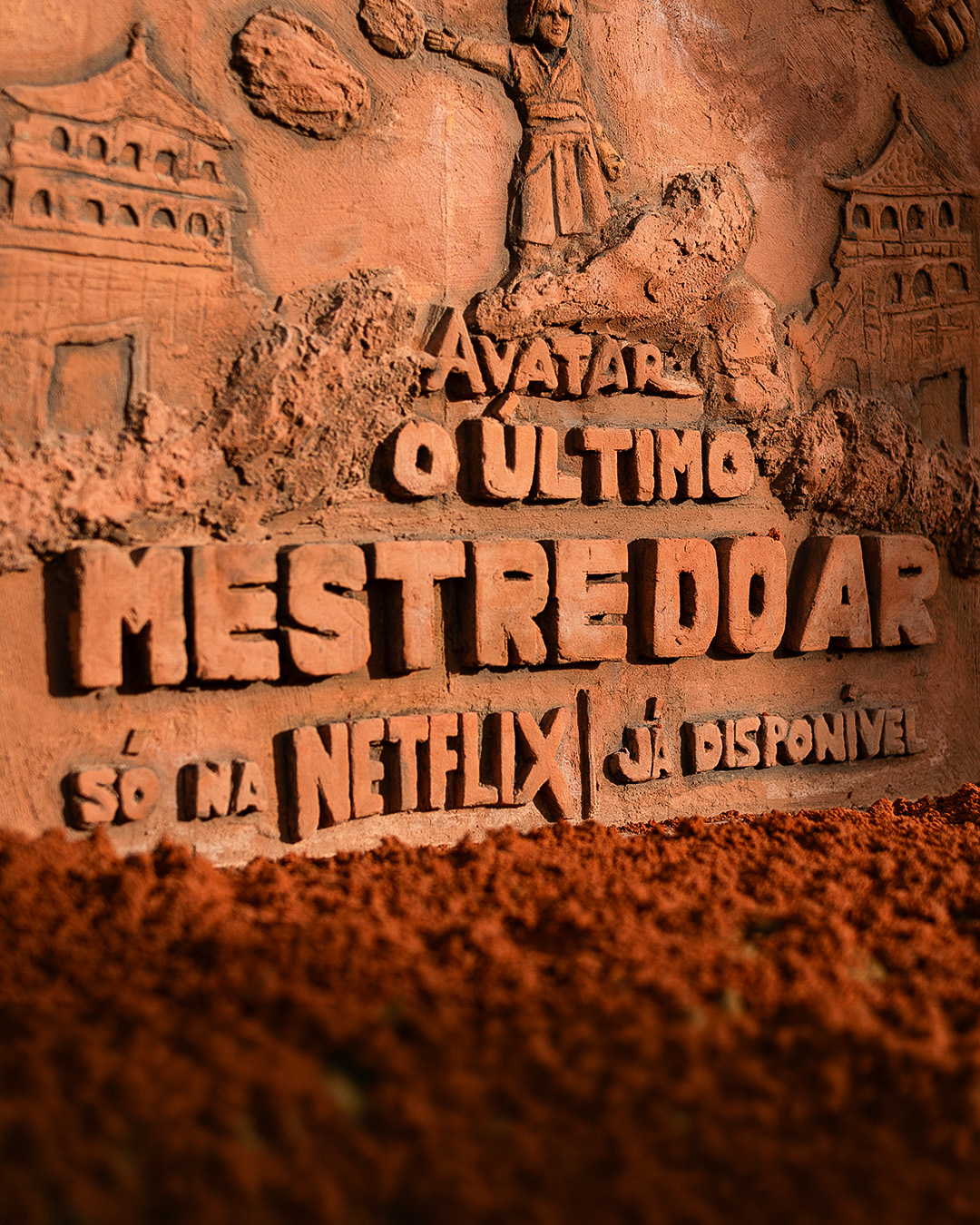 Detail of a clay relief spelling "Avatar: O Último Mestre do Ar" and the Netflix logo above a mound of red earth