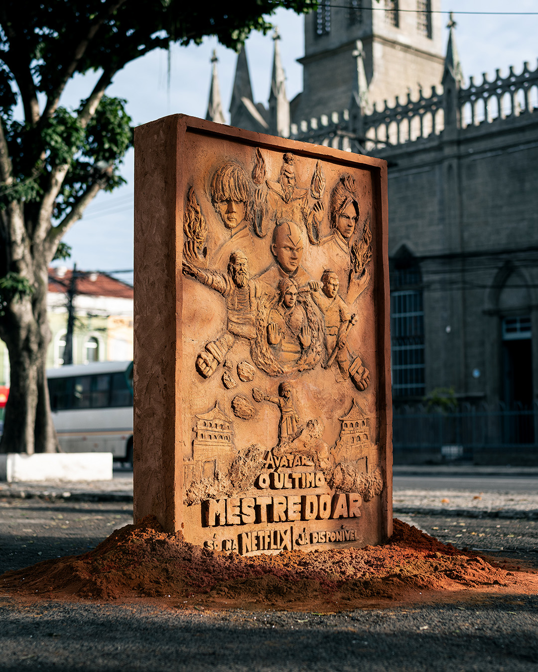 Clay relief poster for "Avatar: O Último Mestre do Ar" set on red earth in a street in front of an old church