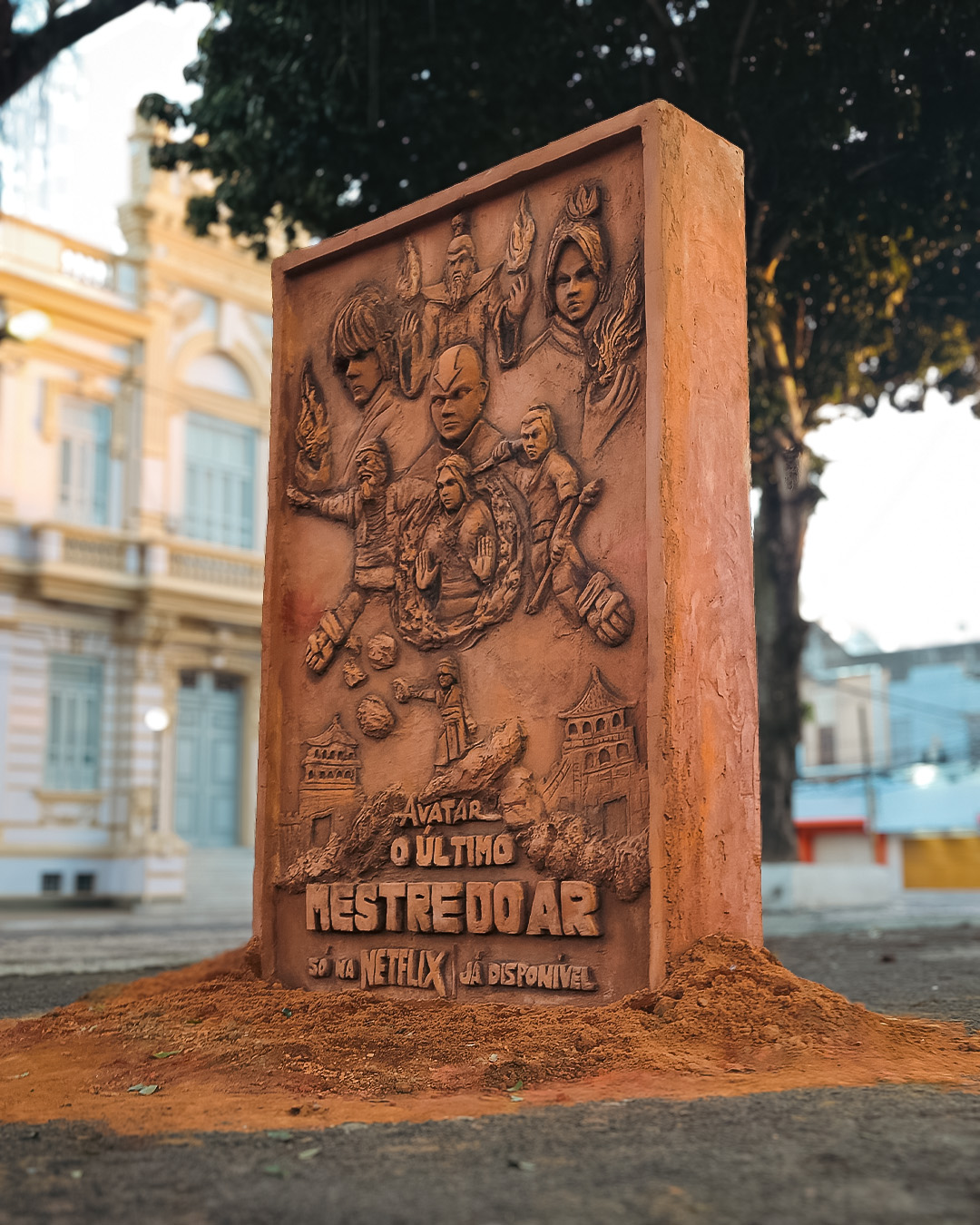 Terracotta relief of Avatar characters on a pile of red soil in a town square, an old building and tree behind it