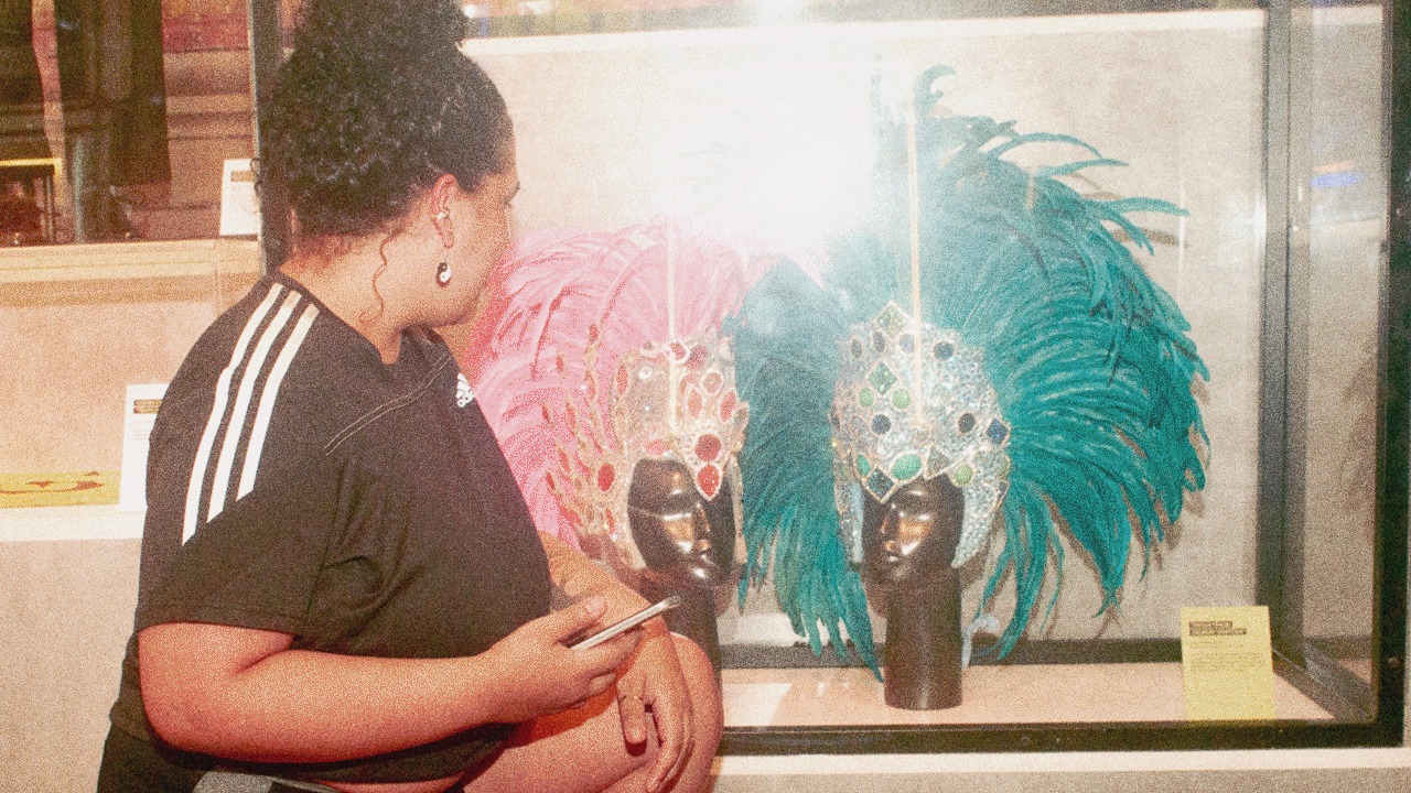 A woman crouches to look at two feathered carnival headdresses in a glass case