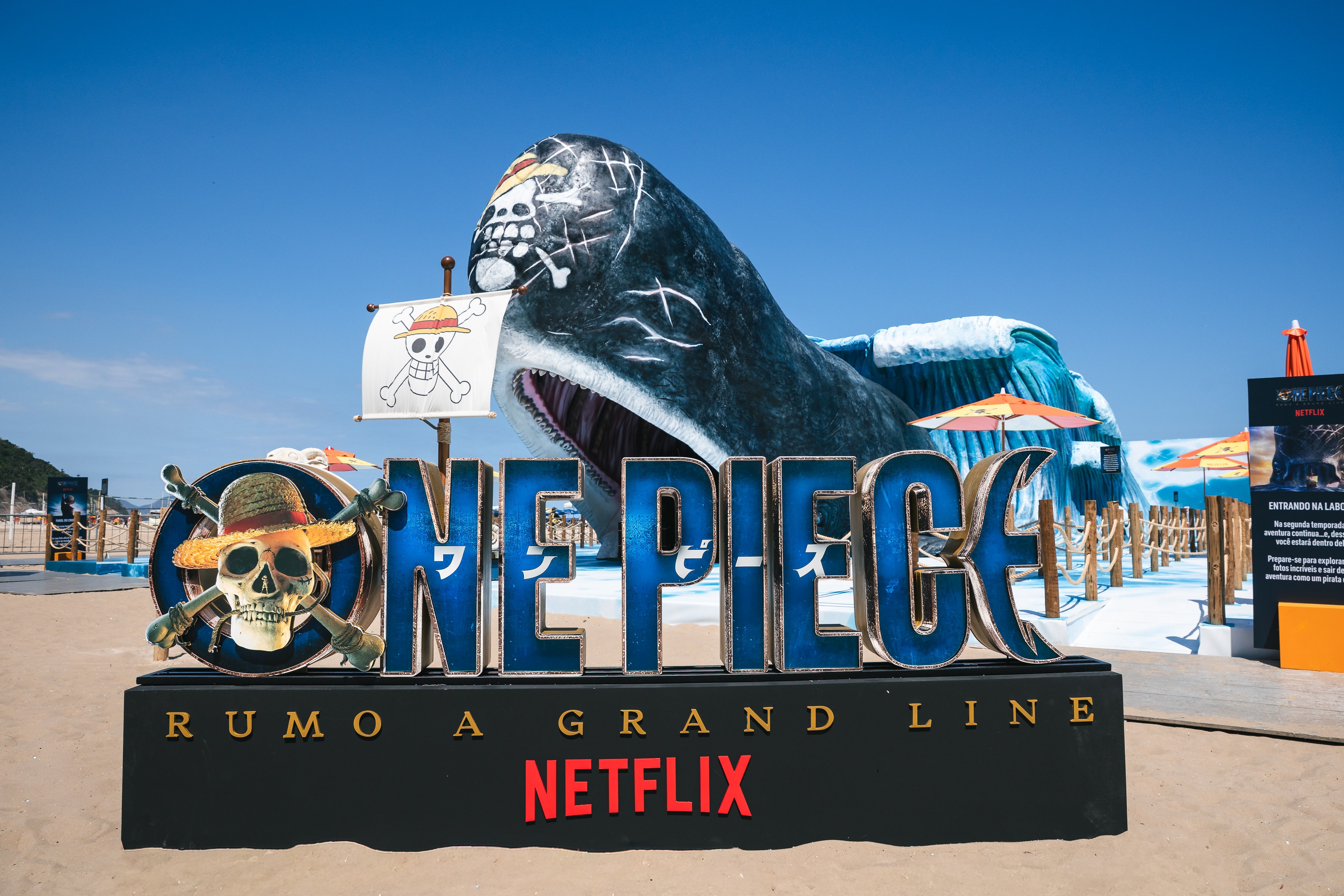 Beach installation with a giant whale sculpture rising behind a large "One Piece" logo and Netflix sign on the sand