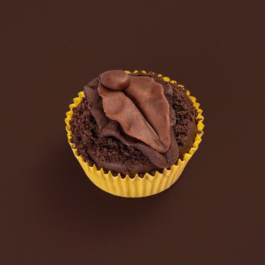 Product shot of a chocolate cupcake with crumb topping and a fondant vulva design in a yellow liner on brown