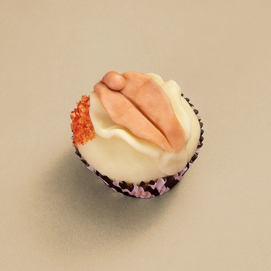 Product shot of a white-frosted cupcake with a peach fondant vulva design and orange crumbs on a beige backdrop