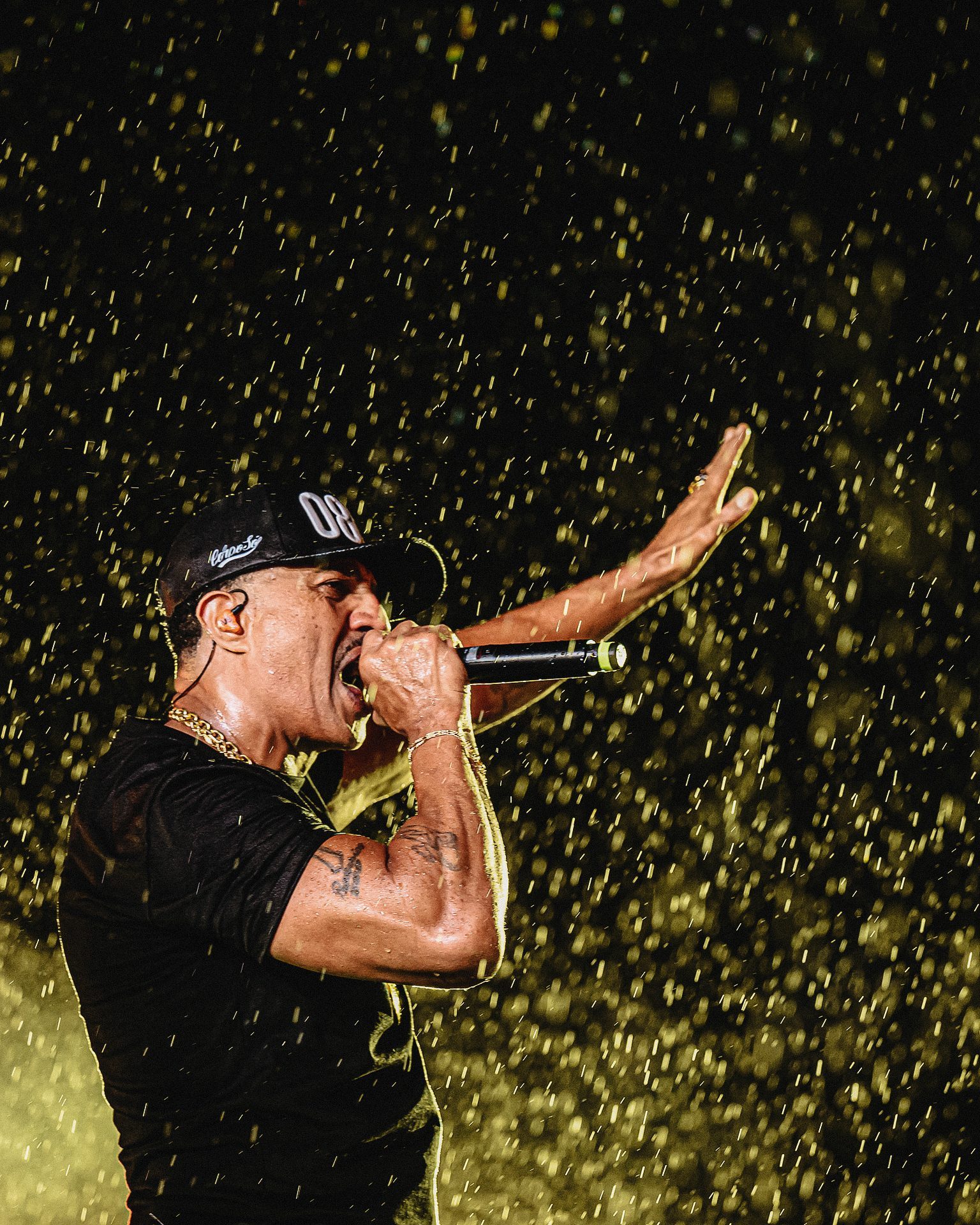 Rapper in a black cap sings into a microphone with one arm outstretched as water spray glitters around him on stage