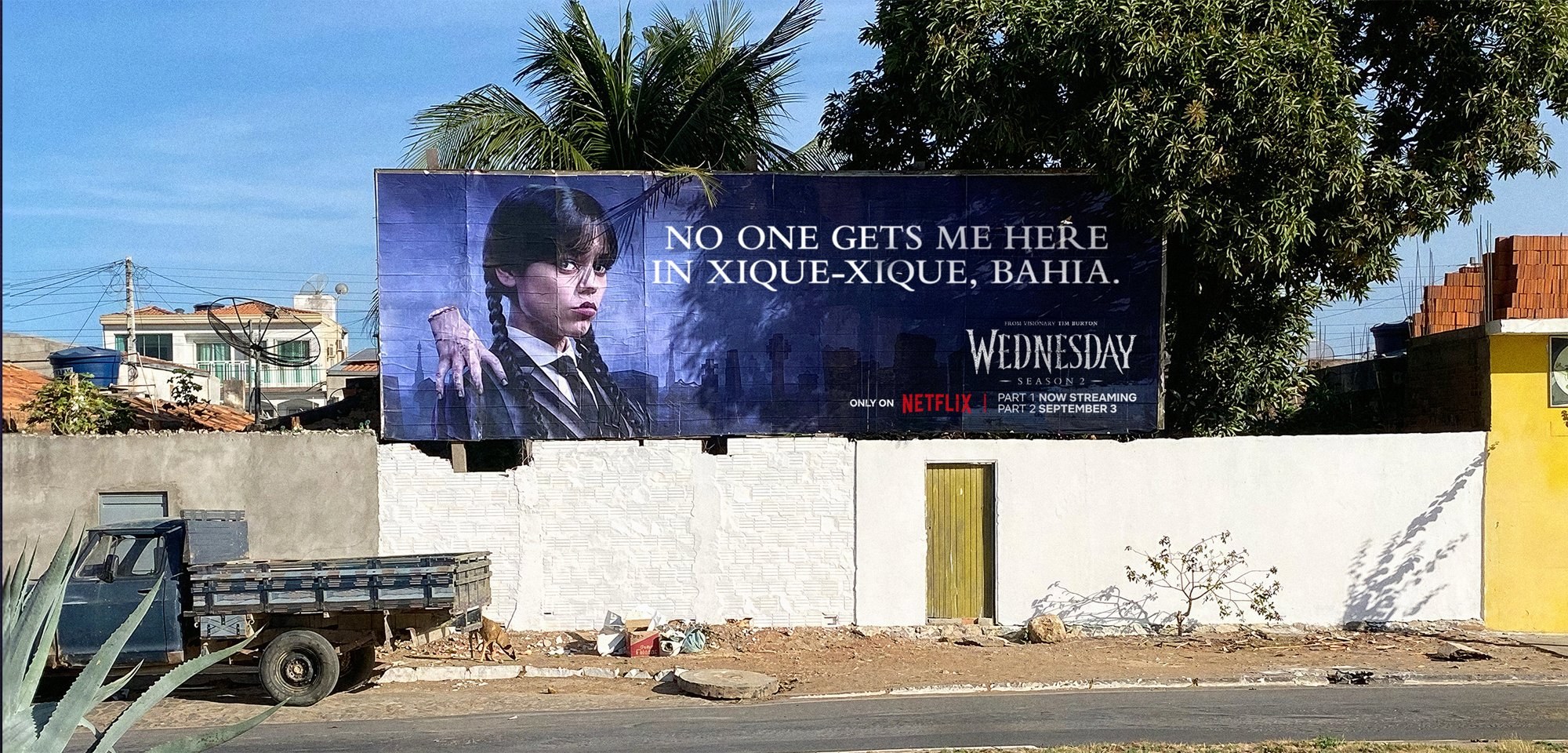 Wednesday Season 2 billboard reading "No one gets me here in Xique-Xique, Bahia" above a white wall on a quiet street