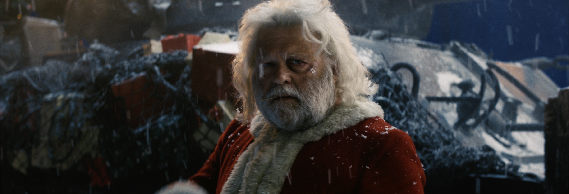 Film still of a white-bearded man in a red Santa coat in falling snow, piled gifts and a snowy tank behind him
