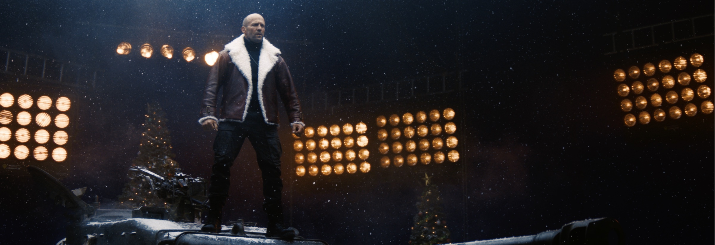 Film still of a man in a shearling jacket standing on a tank at night, framed by banks of stadium lights and snow