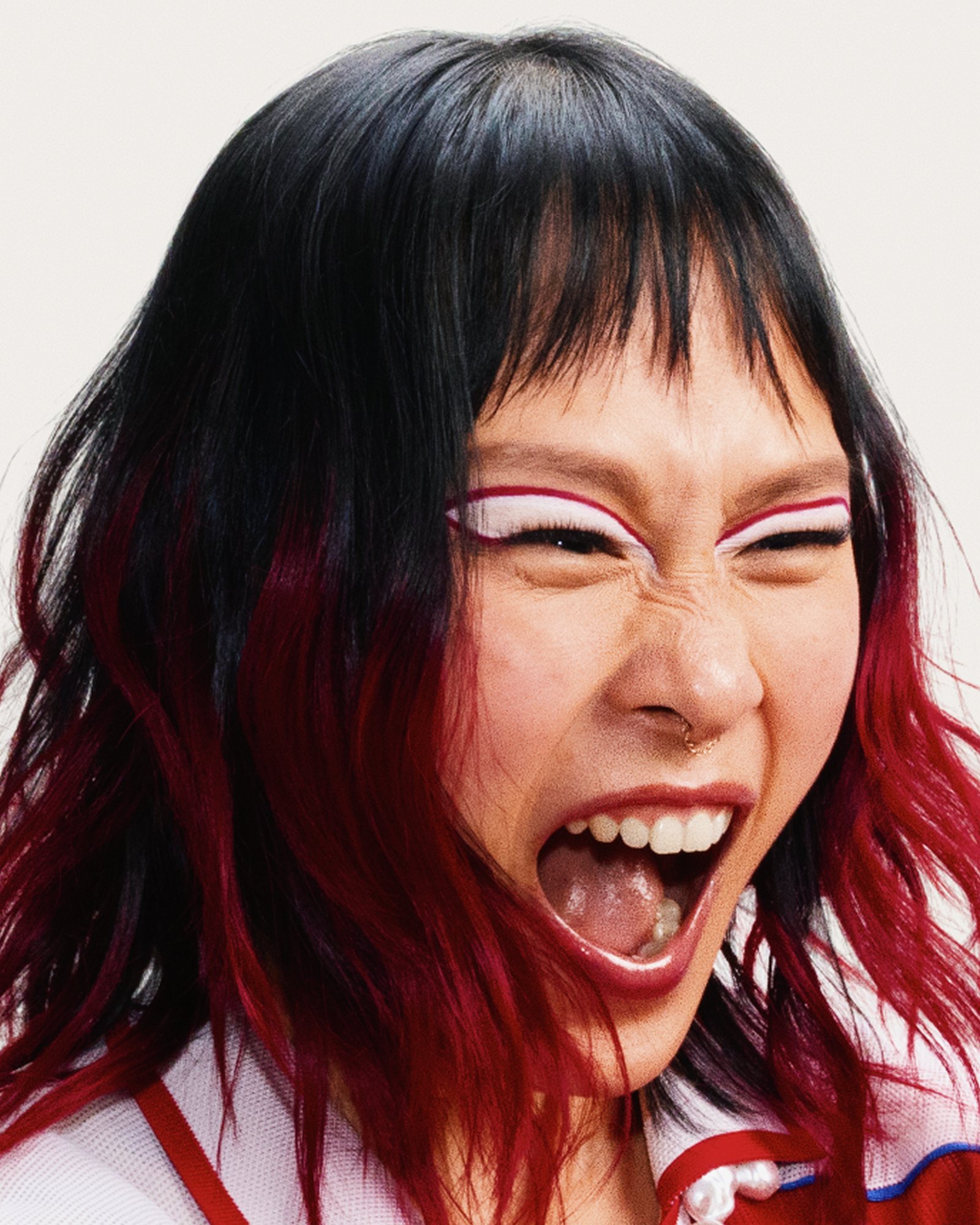 Close-up portrait of a woman with red-tipped hair and graphic white eyeliner shouting, on a white backdrop