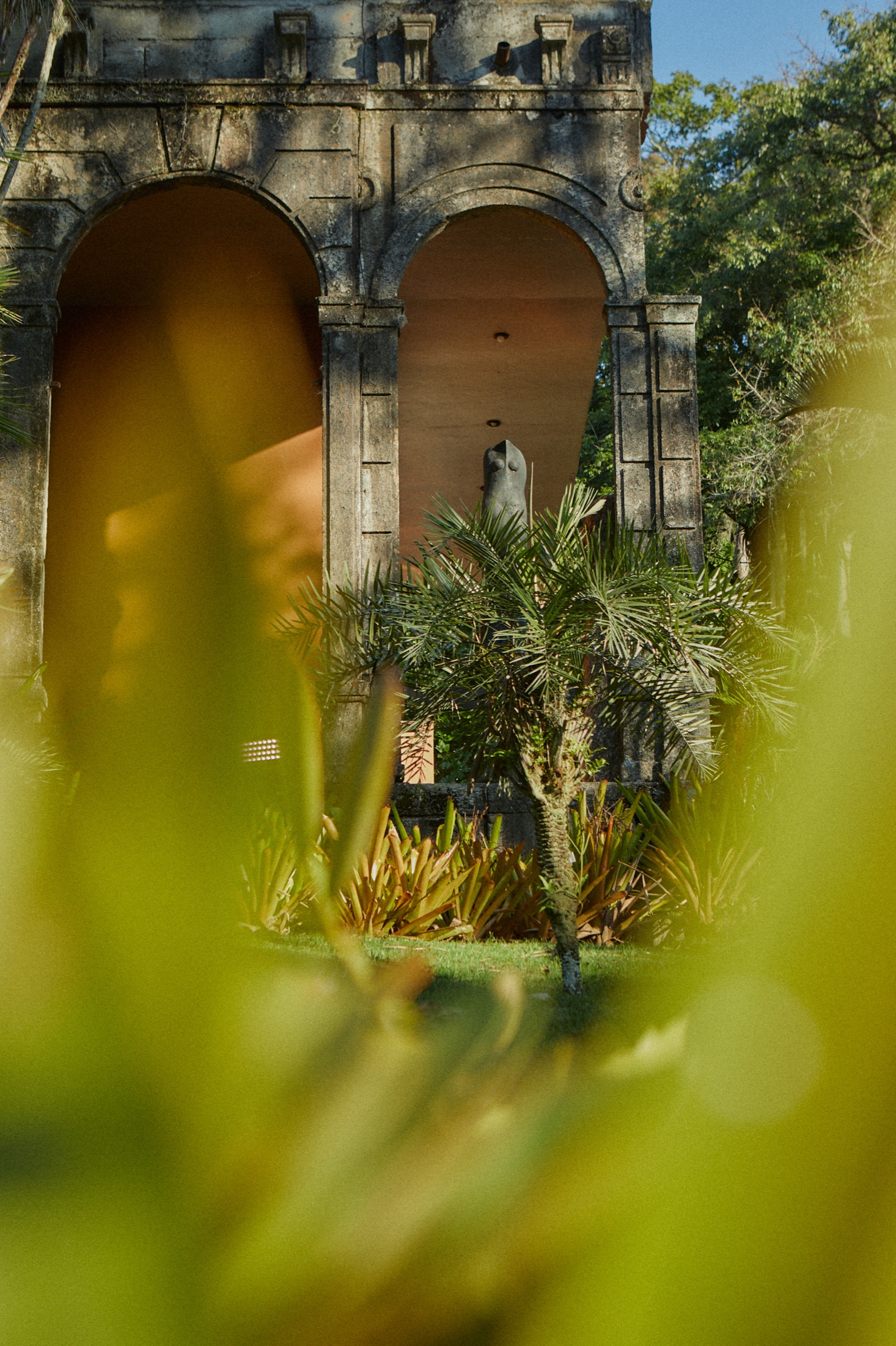 Stone arched ruin with a statue inside, framed by palms and blurred foliage in the foreground