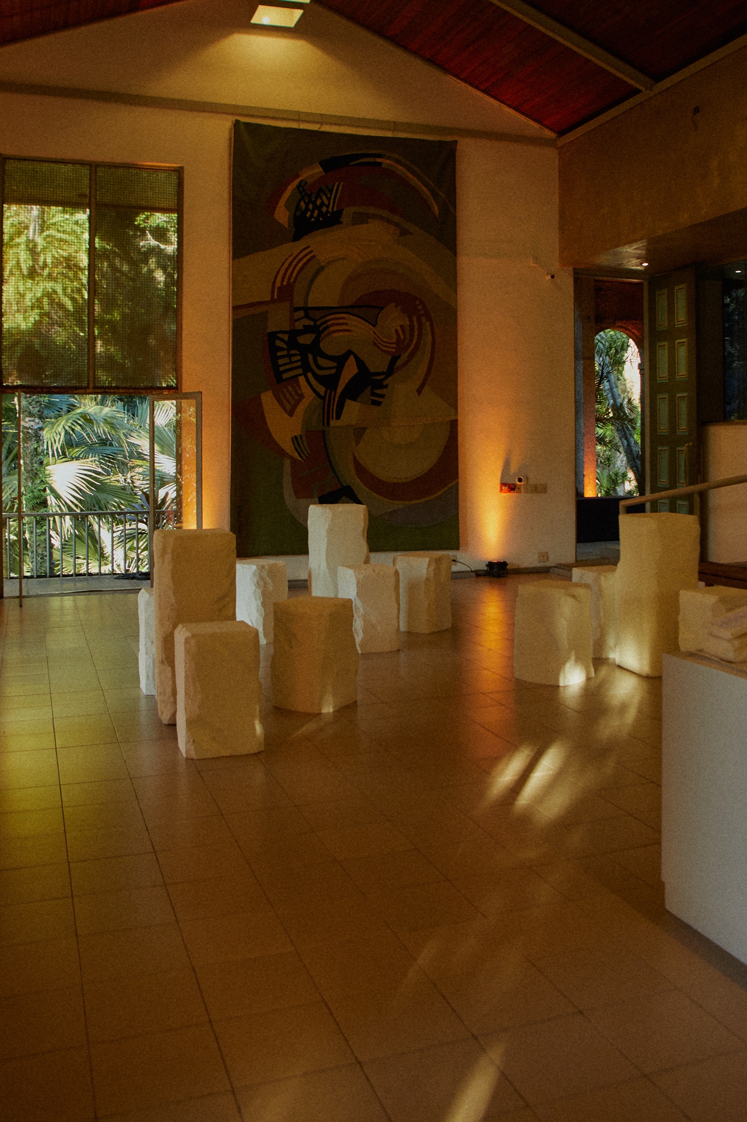 Sunlit hall with a large abstract tapestry on the wall and white stone blocks set out as seats