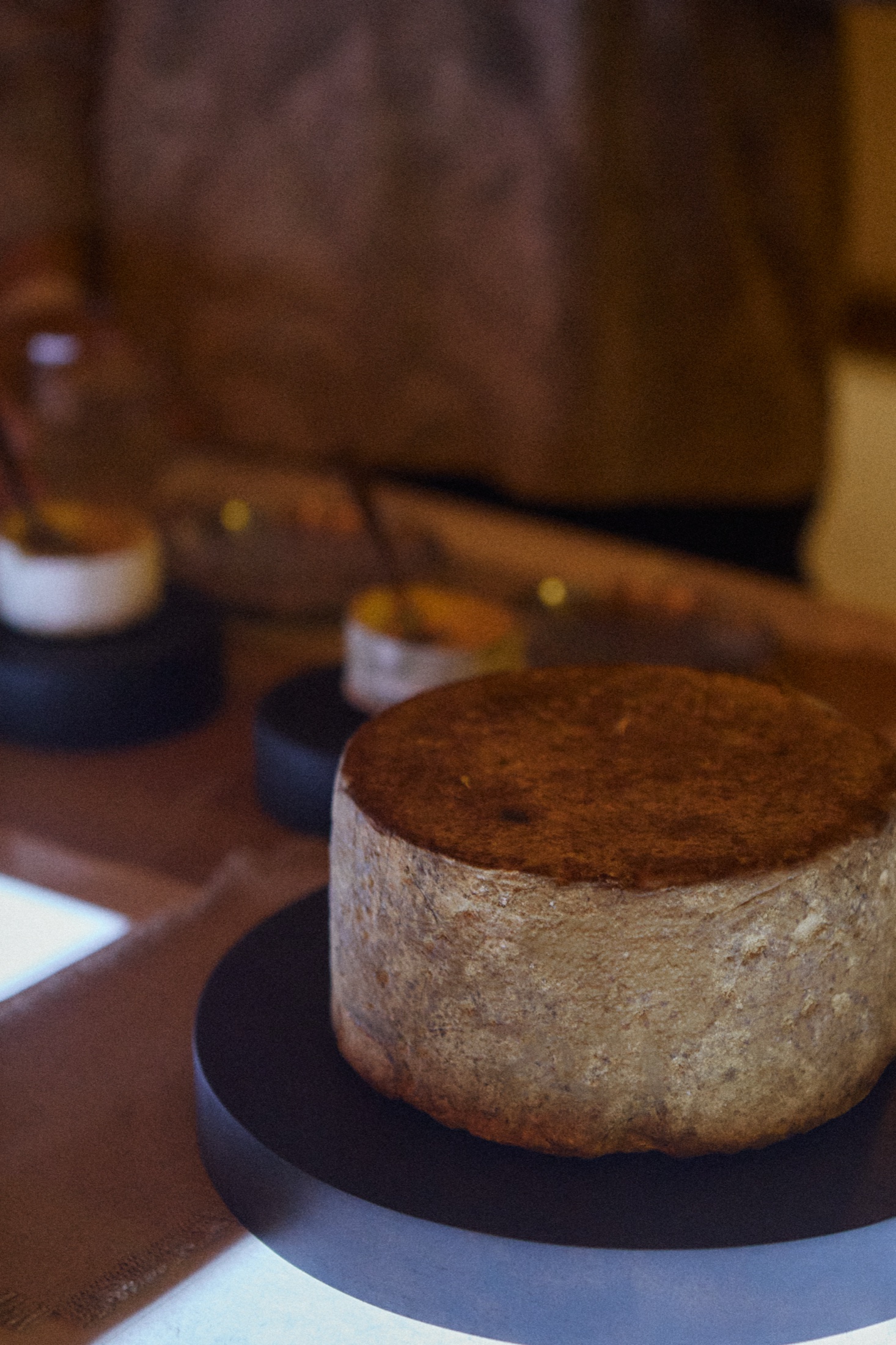 Wheel of aged cheese on a dark round board, with small bowls and a blurred person behind