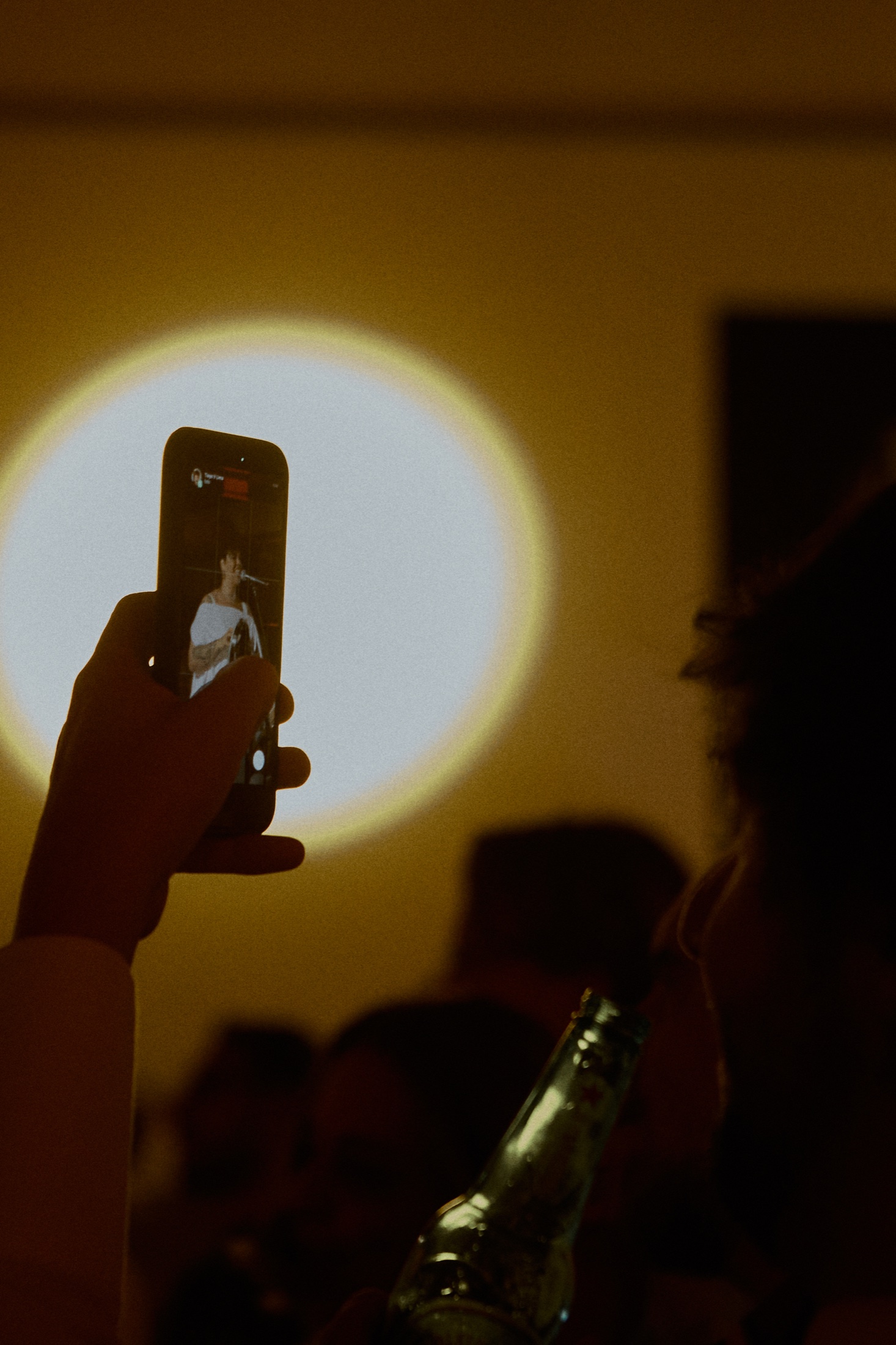 Hand holds up a phone filming a singer, against a circle of projected light at a dim event
