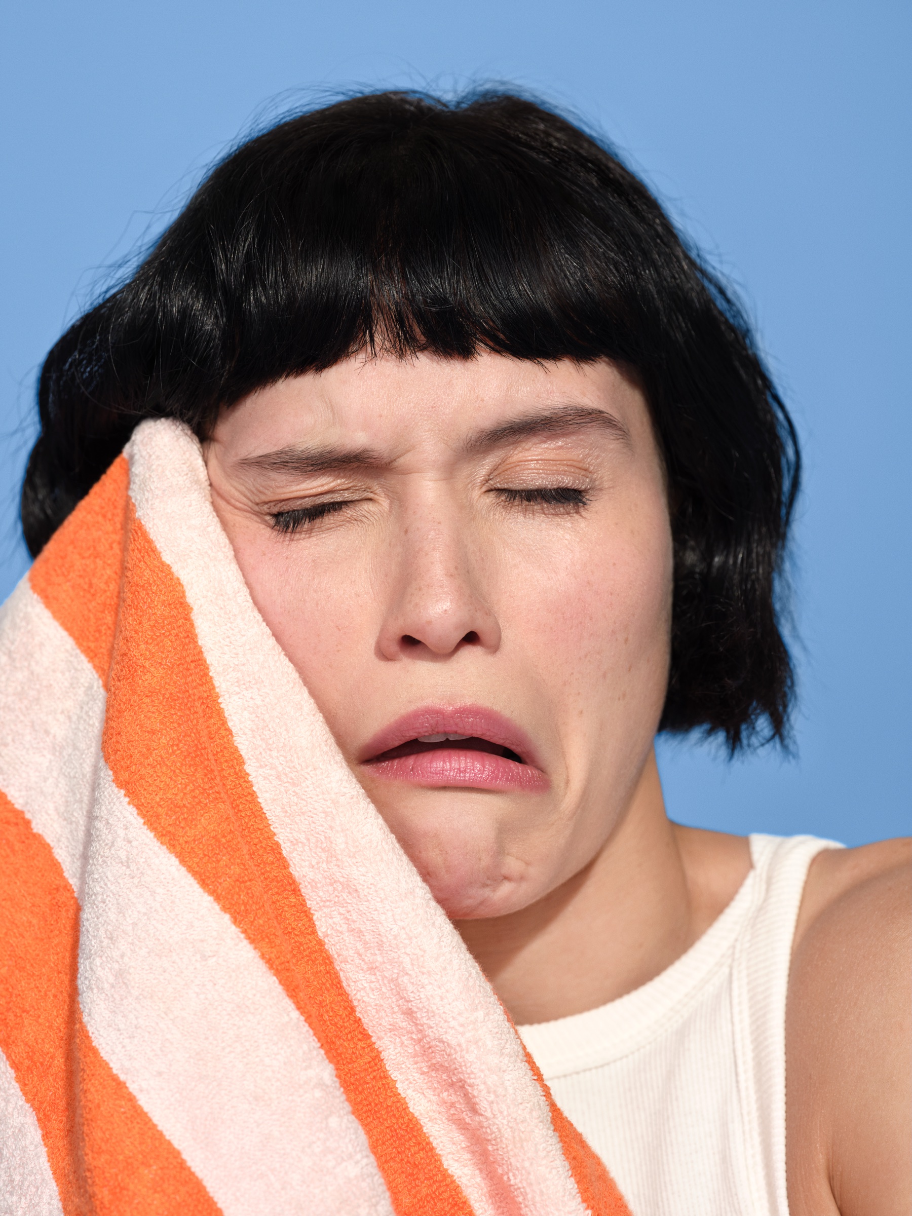 Woman with a black bob grimaces, eyes shut, as she presses an orange and white striped bath towel to her face against a blue backdrop