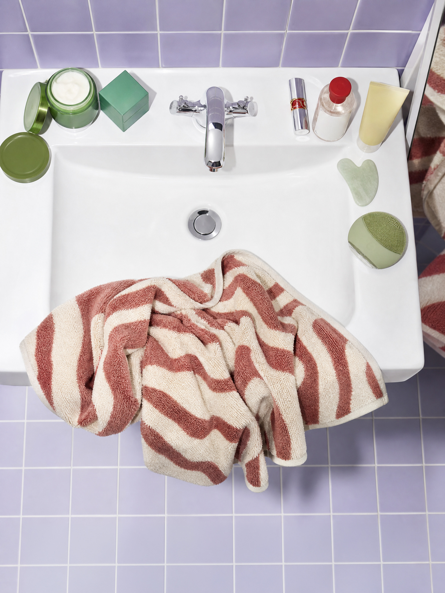 Overhead view of a white bathroom sink with skincare jars and tools on the rim and a crumpled pink striped towel draped over the edge