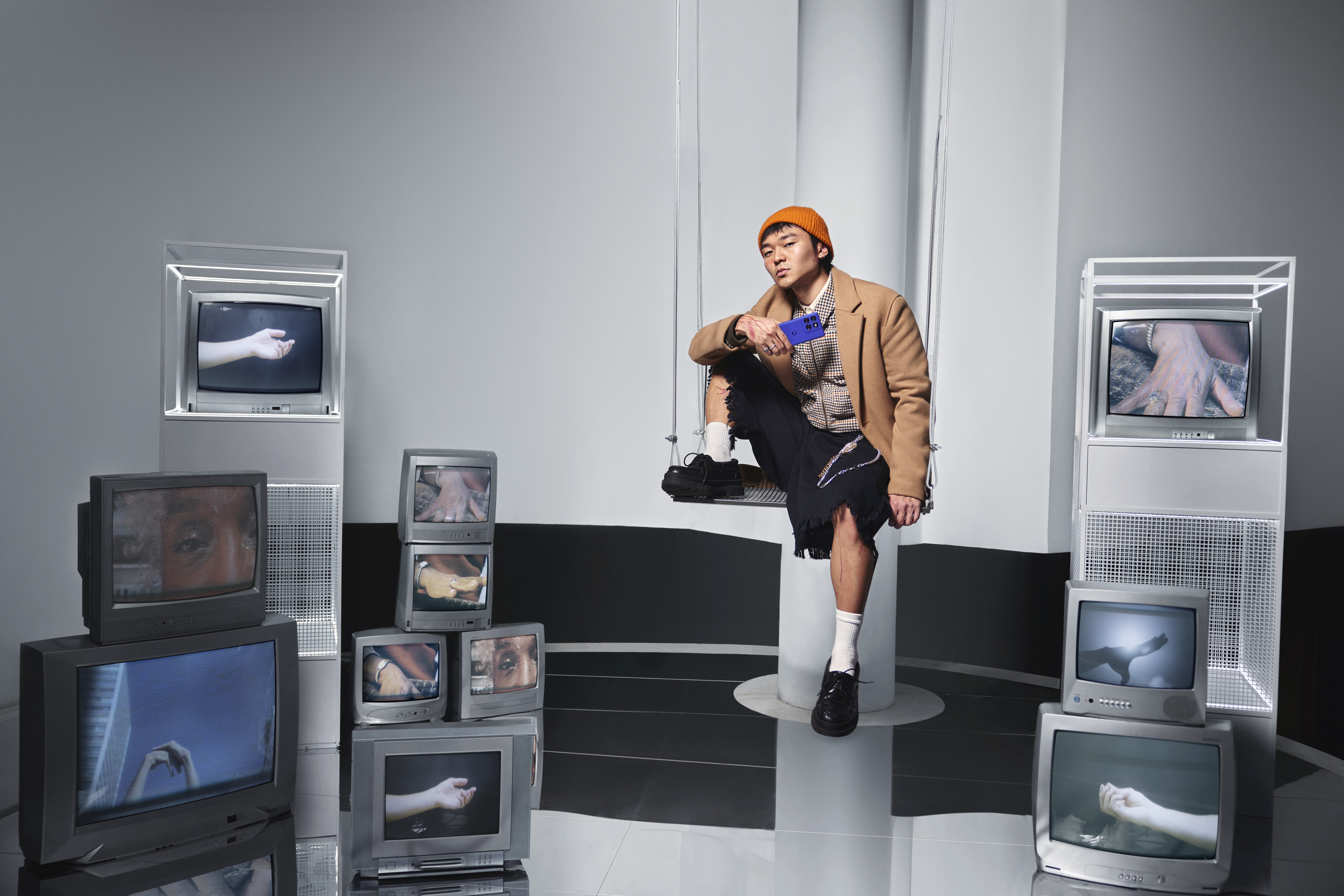Man in an orange beanie sits on a swing holding a blue phone, surrounded by CRT TVs showing hands