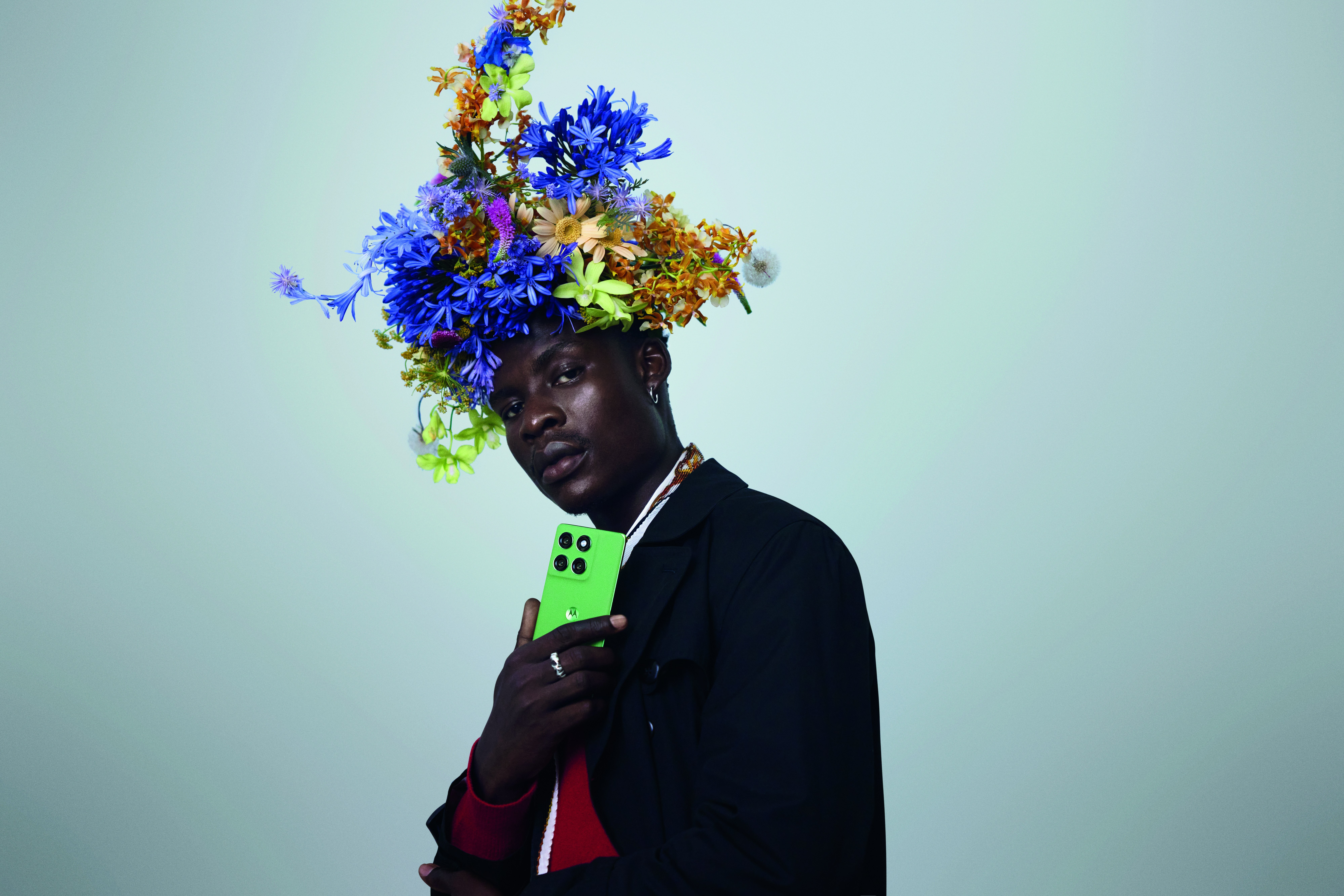 Man wearing a headdress of wildflowers holds a green motorola phone to his chest against a blue sky