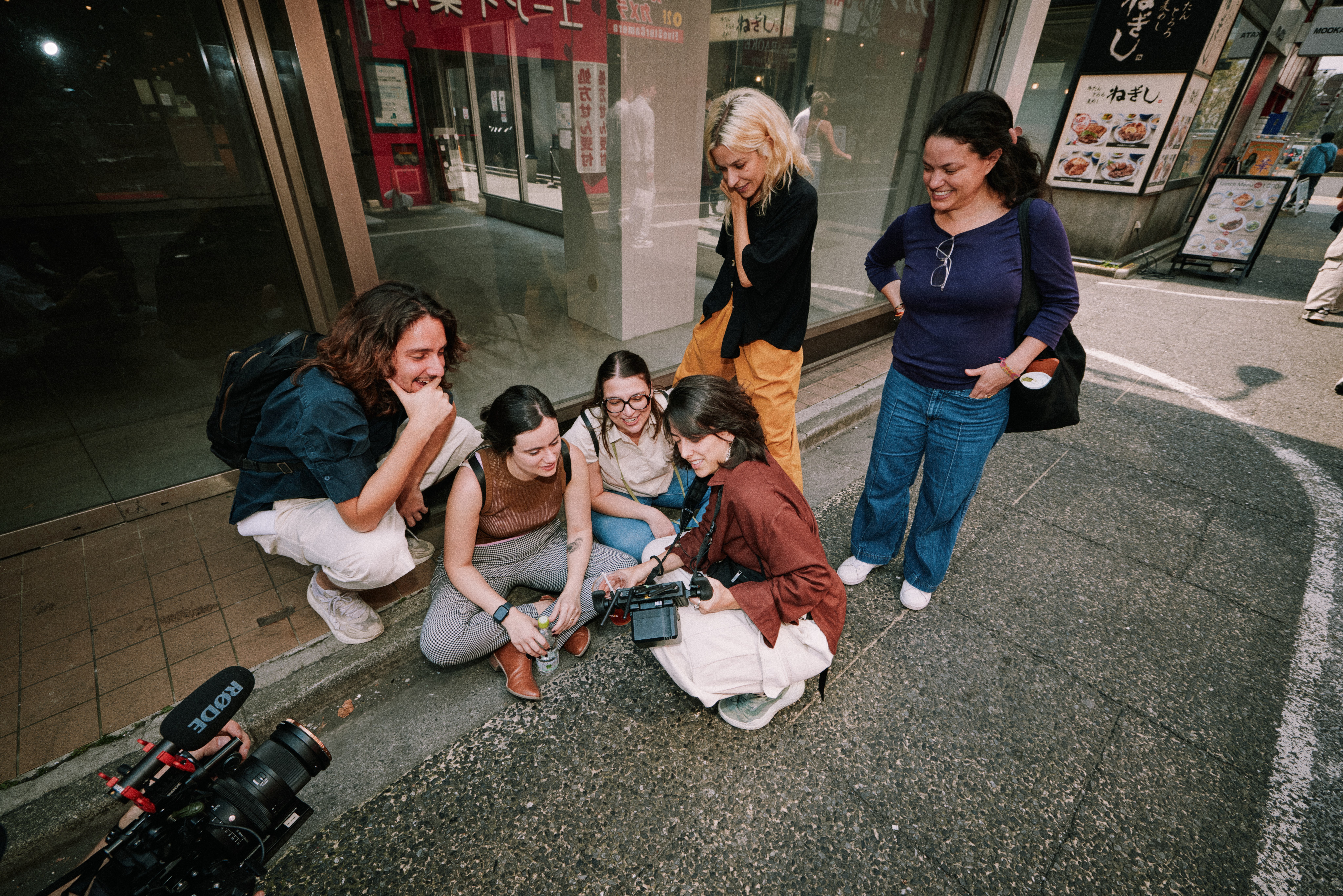 Film crew crouches on a pavement to review footage on a camera monitor, a Rode mic in the foreground