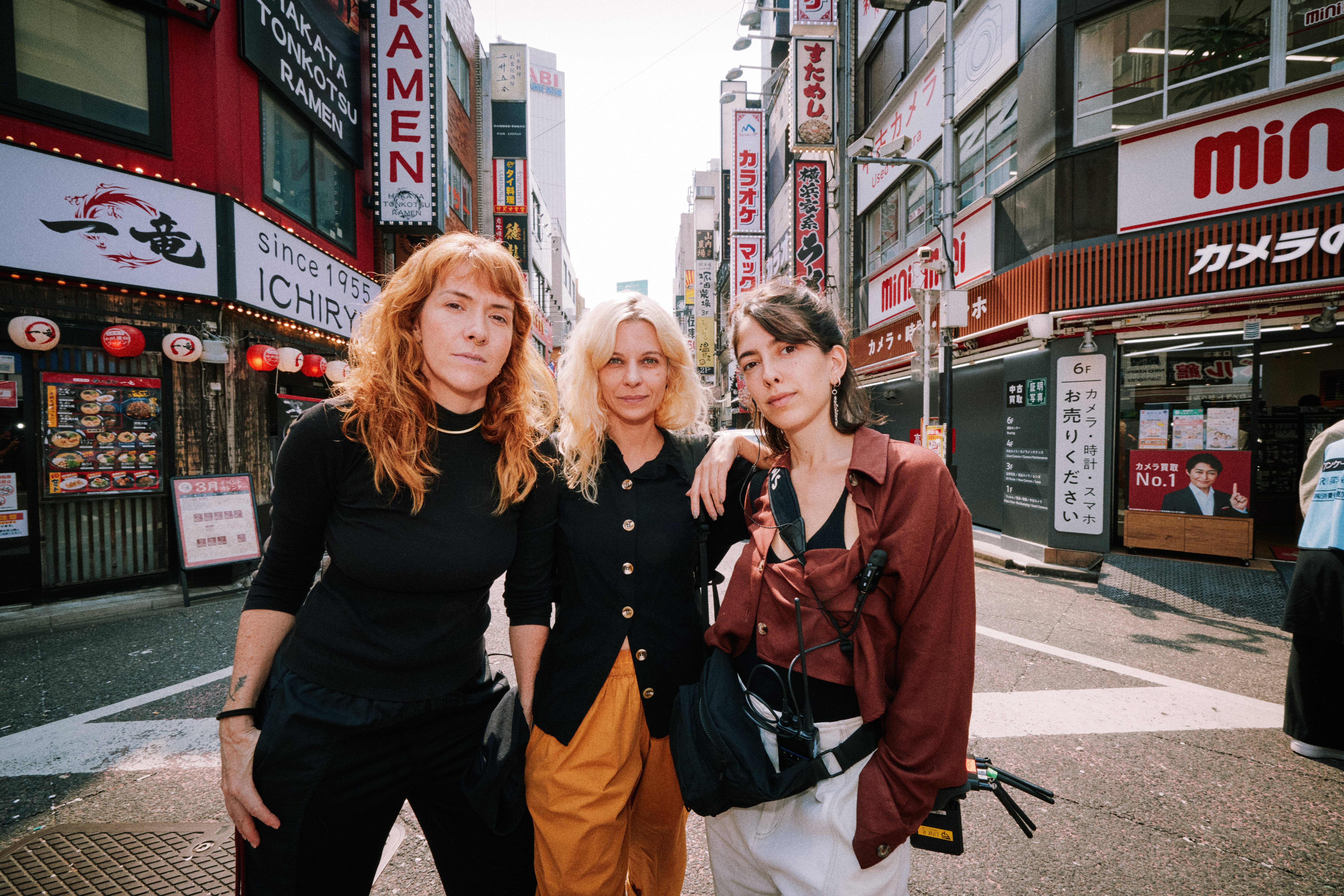 Three women stand together on a busy Japanese street lined with ramen signs and shop fronts