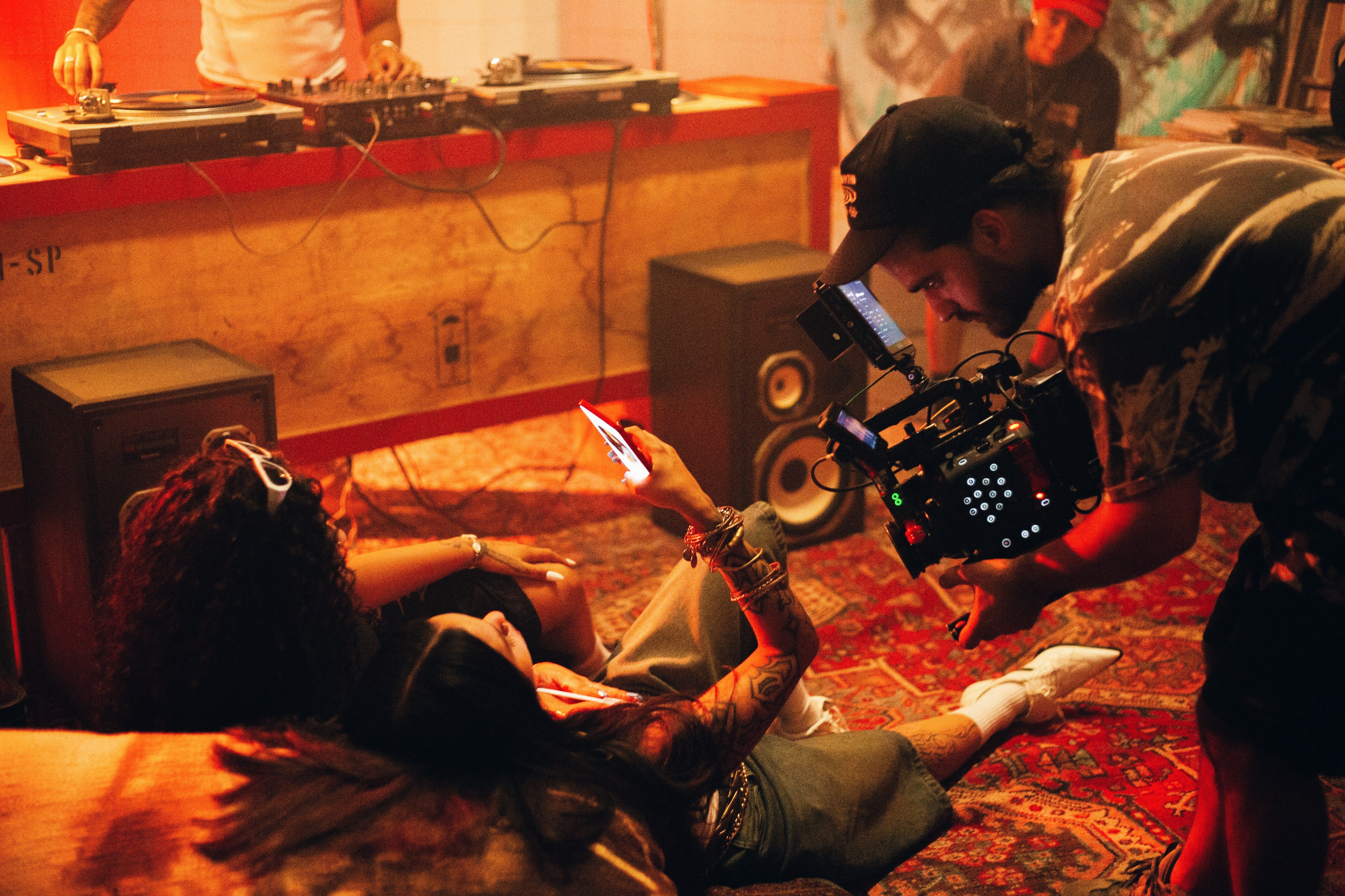 Camera operator films two women lying on a rug, one holding up a phone, while a DJ plays behind them