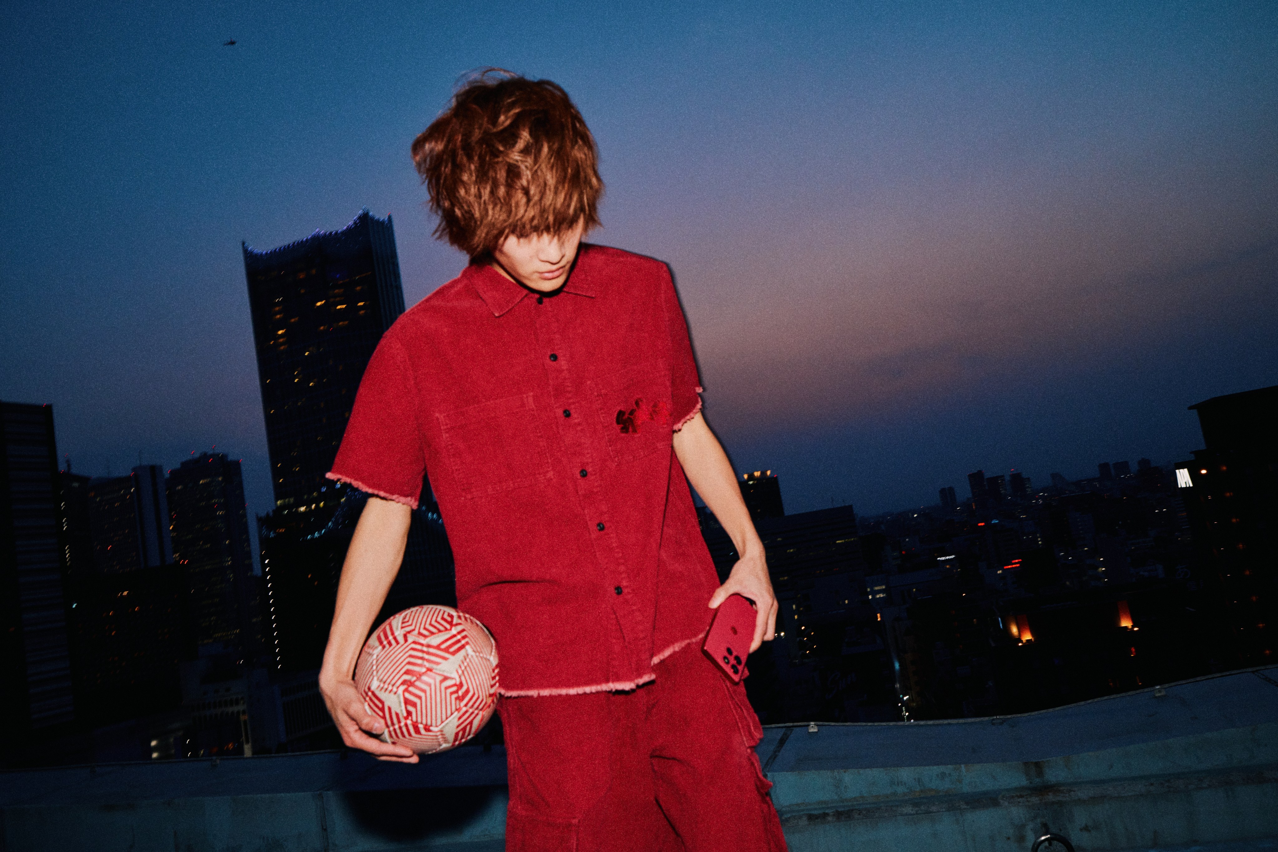 Young man in a red shirt holds a football and a red phone on a rooftop at dusk above the city