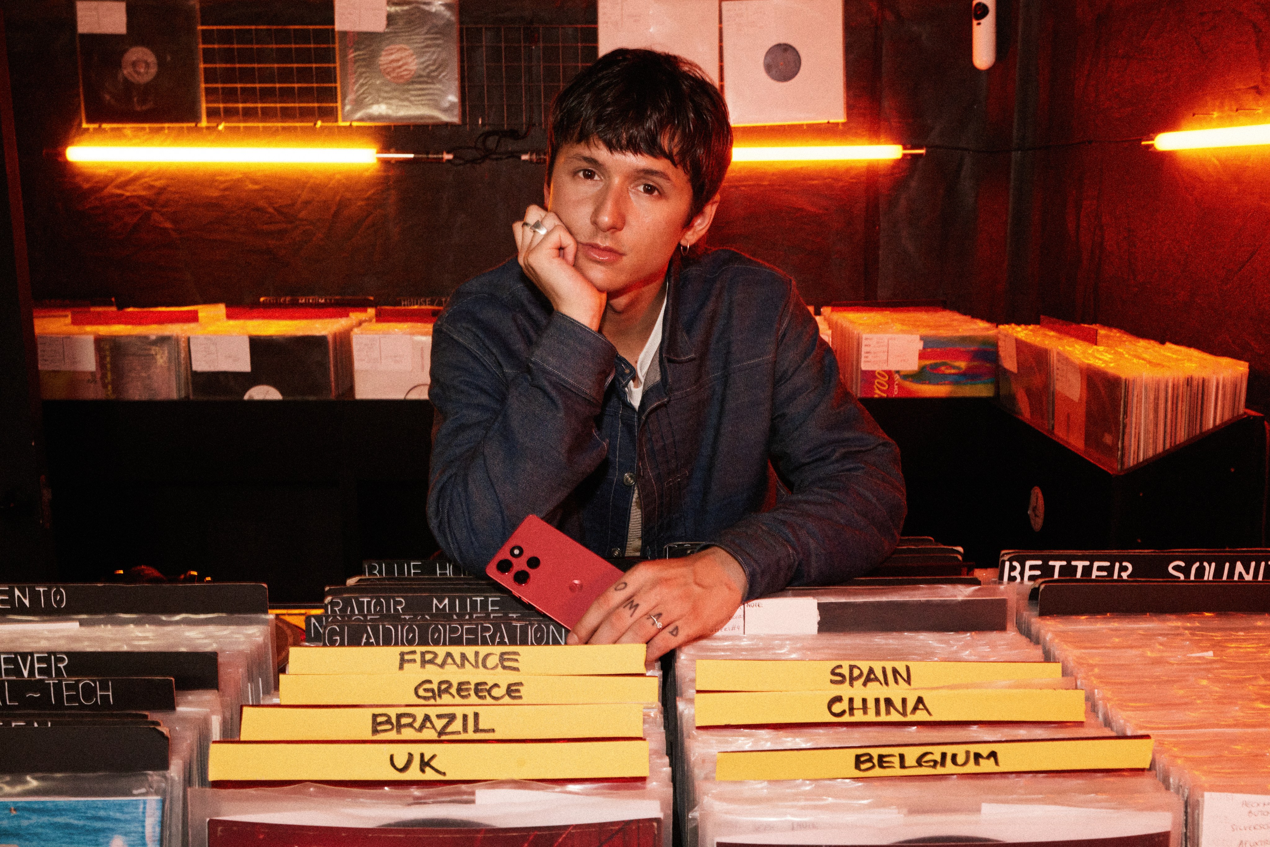 Young man in a denim jacket leans on vinyl record bins holding a red phone, orange neon behind