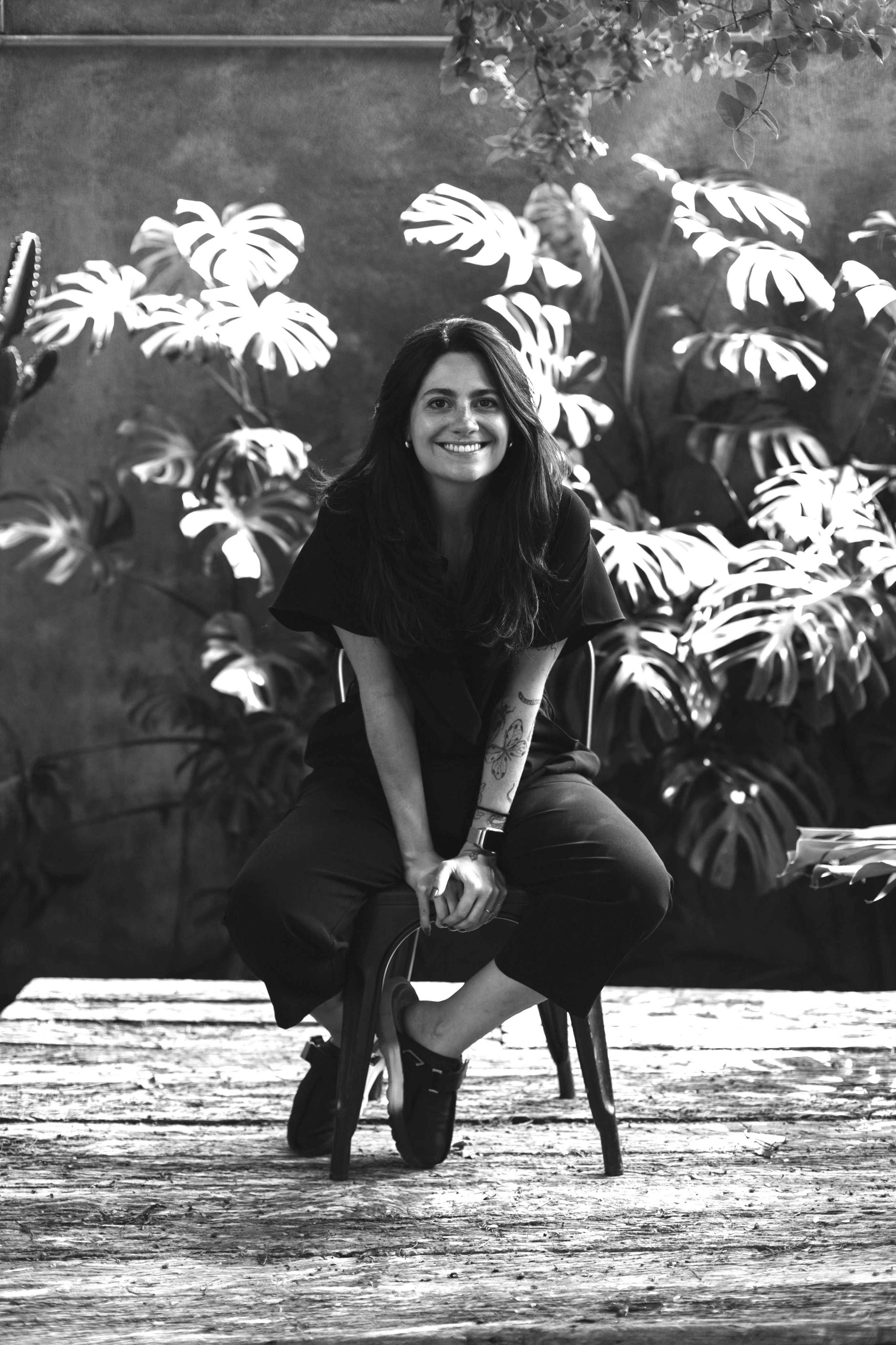 Black and white portrait of a smiling woman seated on a stool in front of large monstera leaves