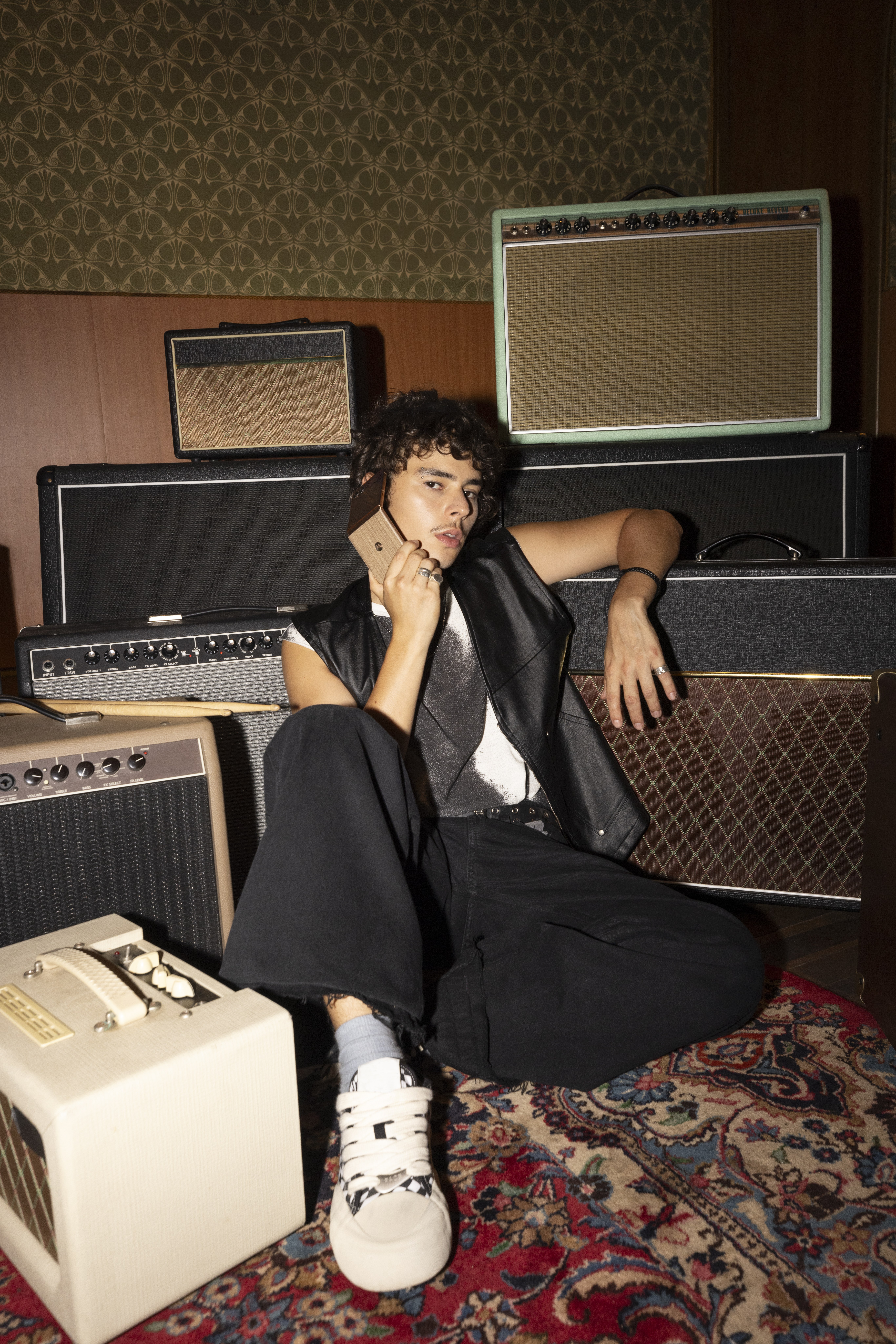 Young man in a leather vest sits on a rug among guitar amps, holding a gold flip phone to his ear
