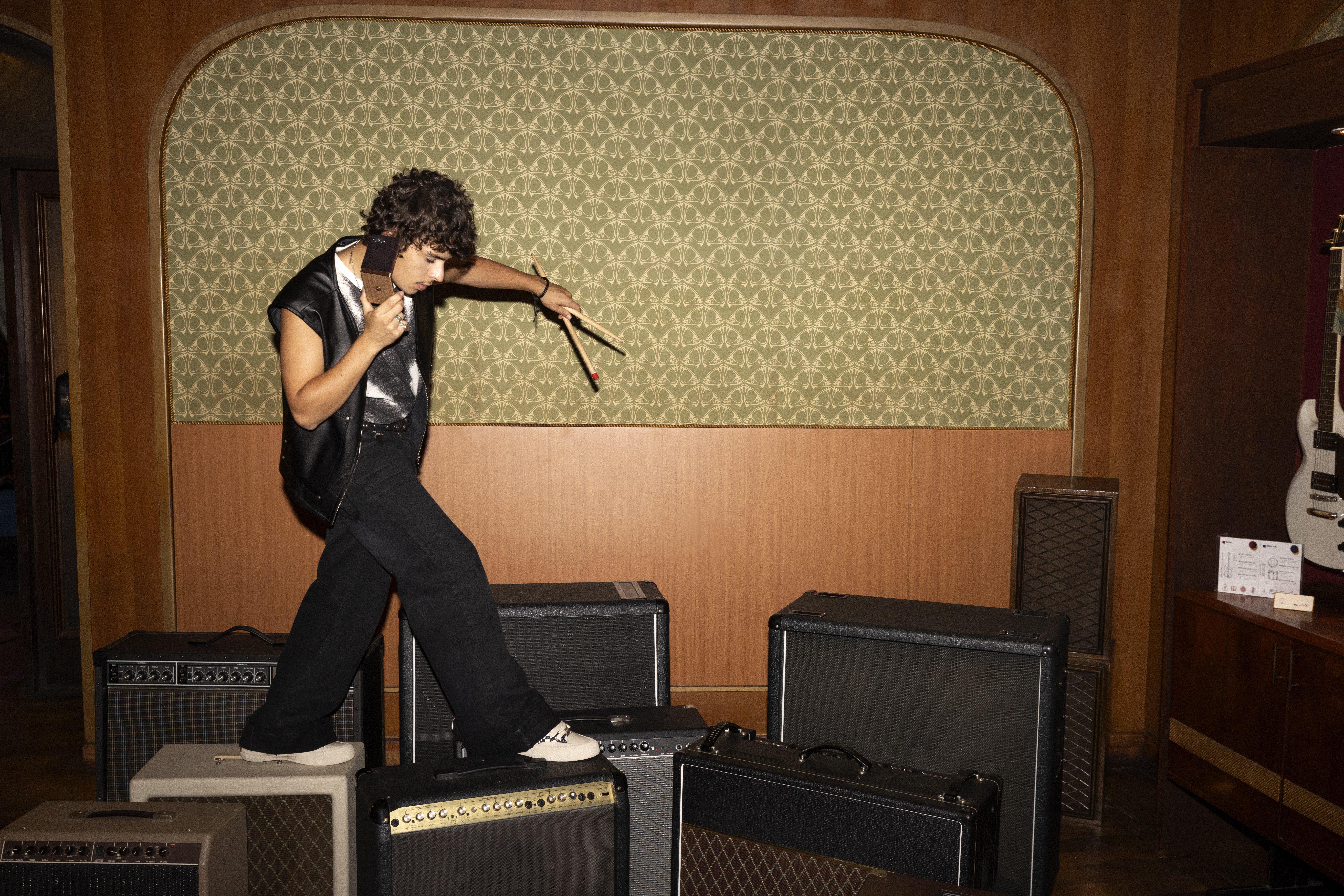 Young man in a leather vest steps across guitar amps, drumsticks in one hand, a phone at his ear