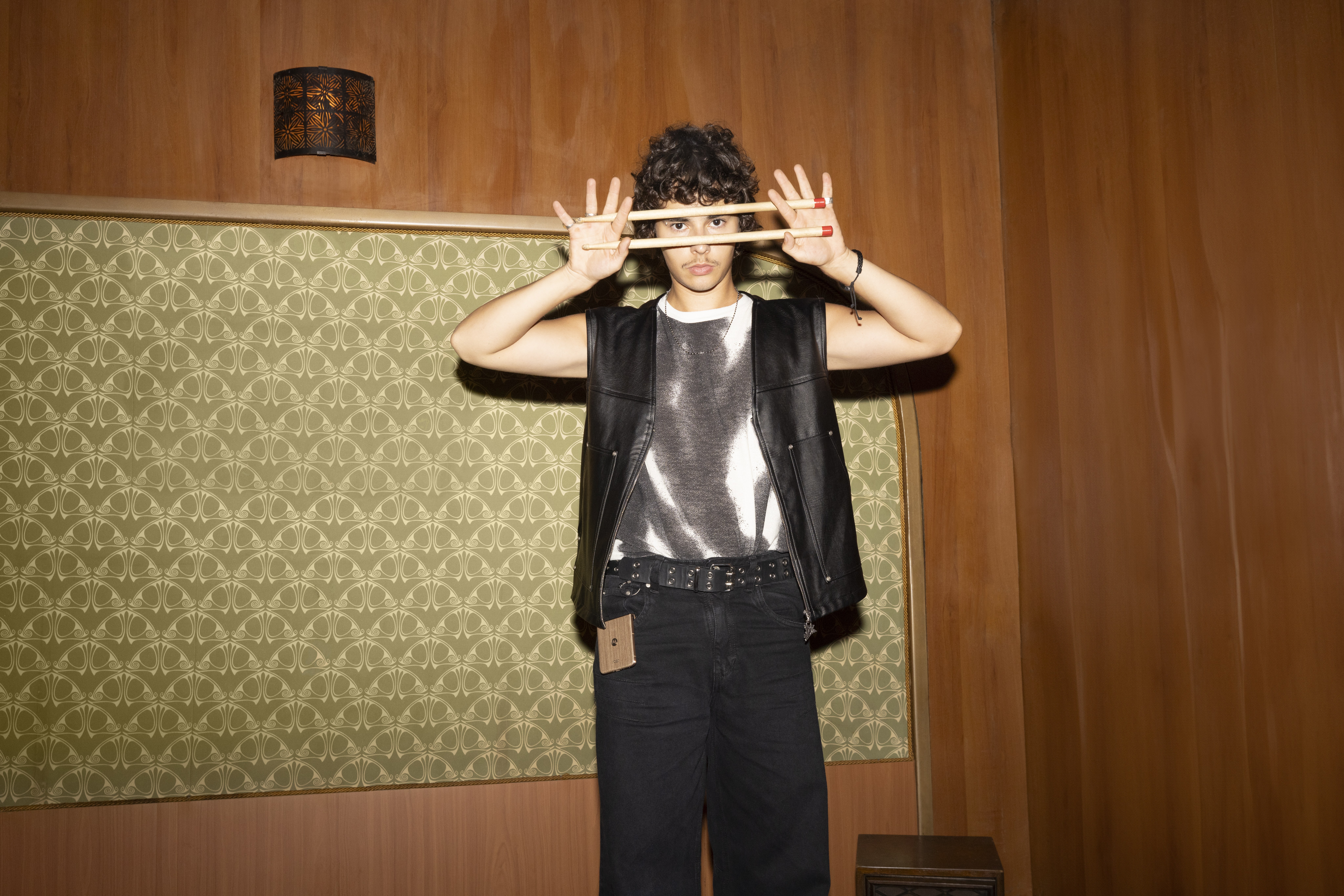 Young man holds two drumsticks across his face, a flip phone clipped to his belt, patterned wallpaper behind